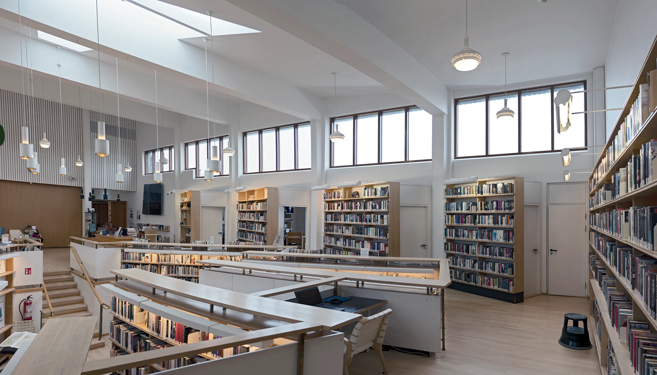 Bright library interior with white bookshelves, reading tables, pendant lights, and large windows along the upper wall