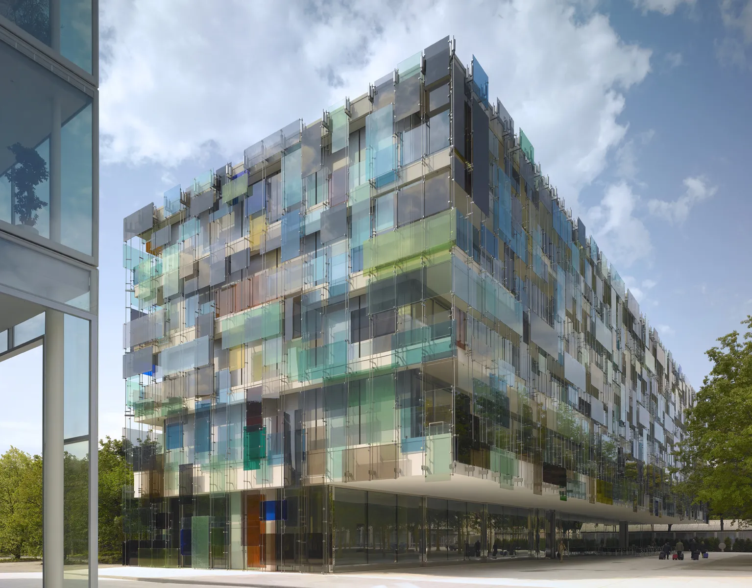 Groot modern gebouw van meerdere verdiepingen met een kleurrijke glazen gevel in blauw- en groentinten onder een halfbewolkte lucht
