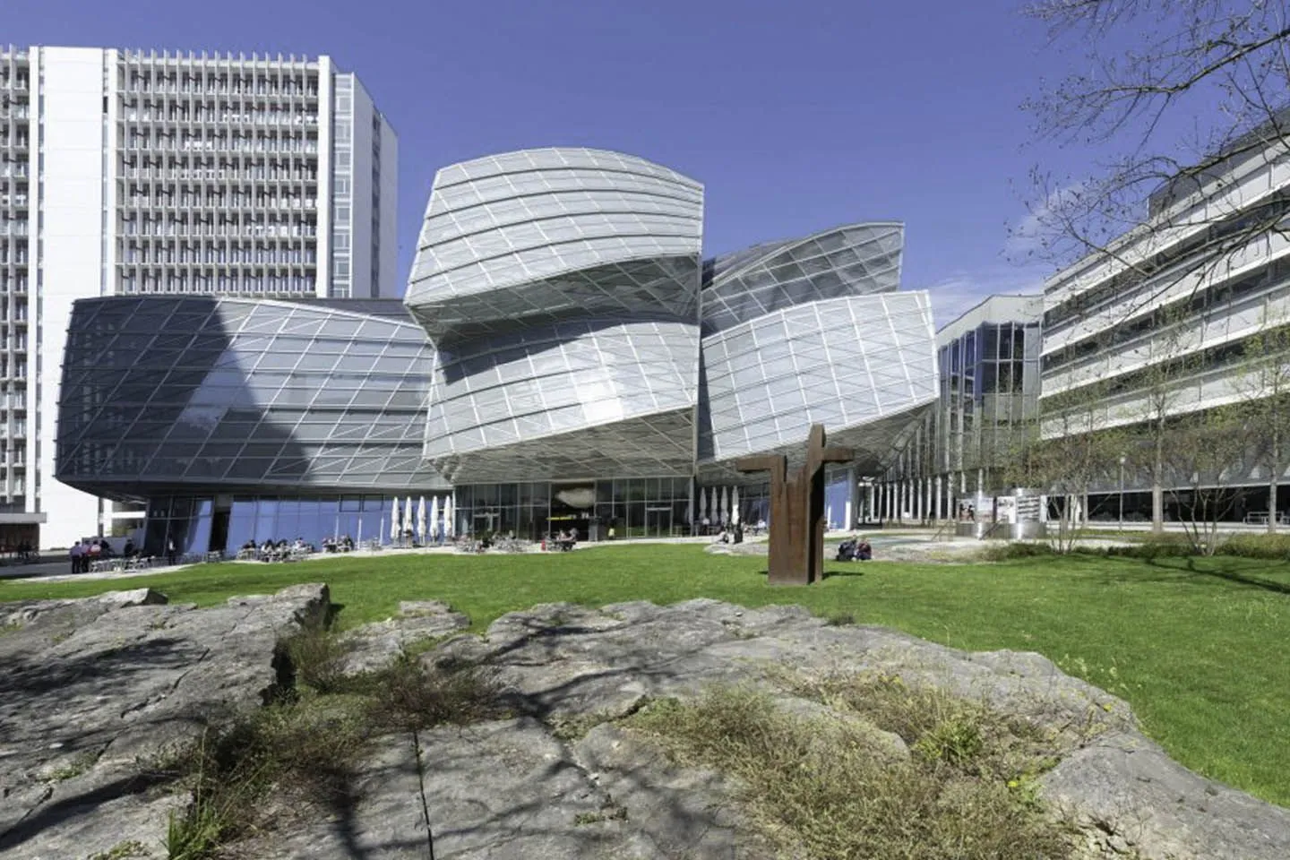 Gebogen gebouw van gelaagd glas en staal met een golvende gevel op de Novartis Campus, geplaatst tegen een groen gazon met rotspartijen op de voorgrond en hoge kantoorgebouwen aan weerszijden onder een helderblauwe lucht.