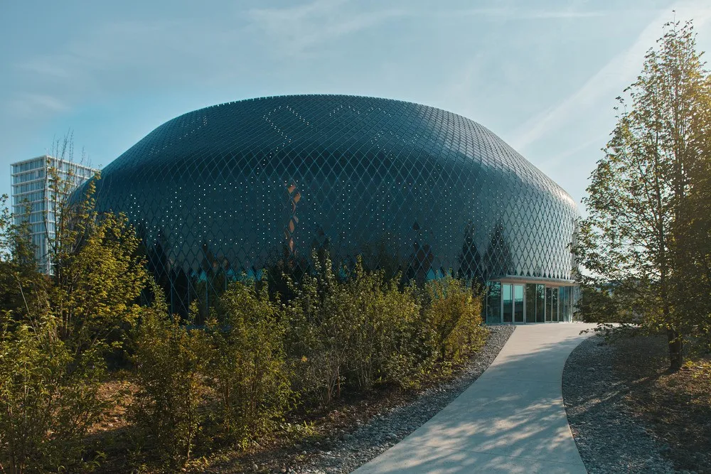 The Novartis Pavillon, a large rounded dome-shaped building with a dark patterned facade, surrounded by trees with a curved pathway leading to the glass entrance