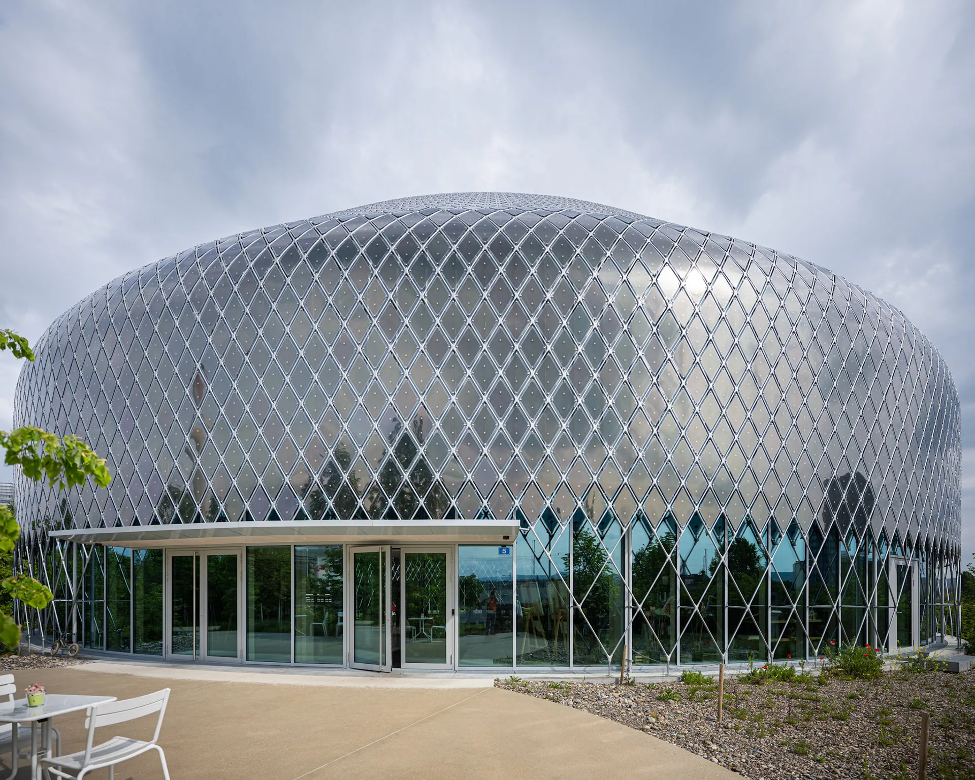 Exterior view of the Novartis Pavillon featuring a large, oval-shaped building with a reflective, silver diamond-patterned facade and glass entrance doors.