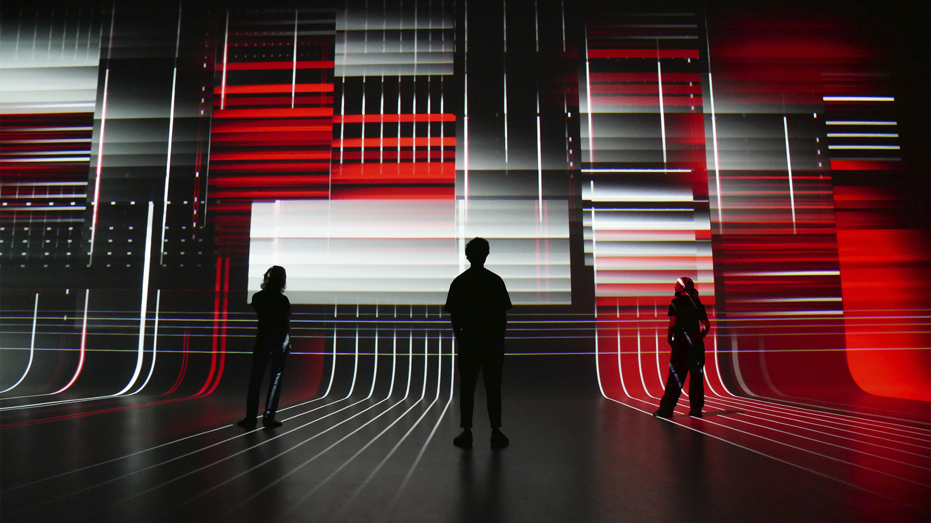 Dark room with red and white digital projections on walls, grid pattern on floor, and three silhouetted figures observing the installation.