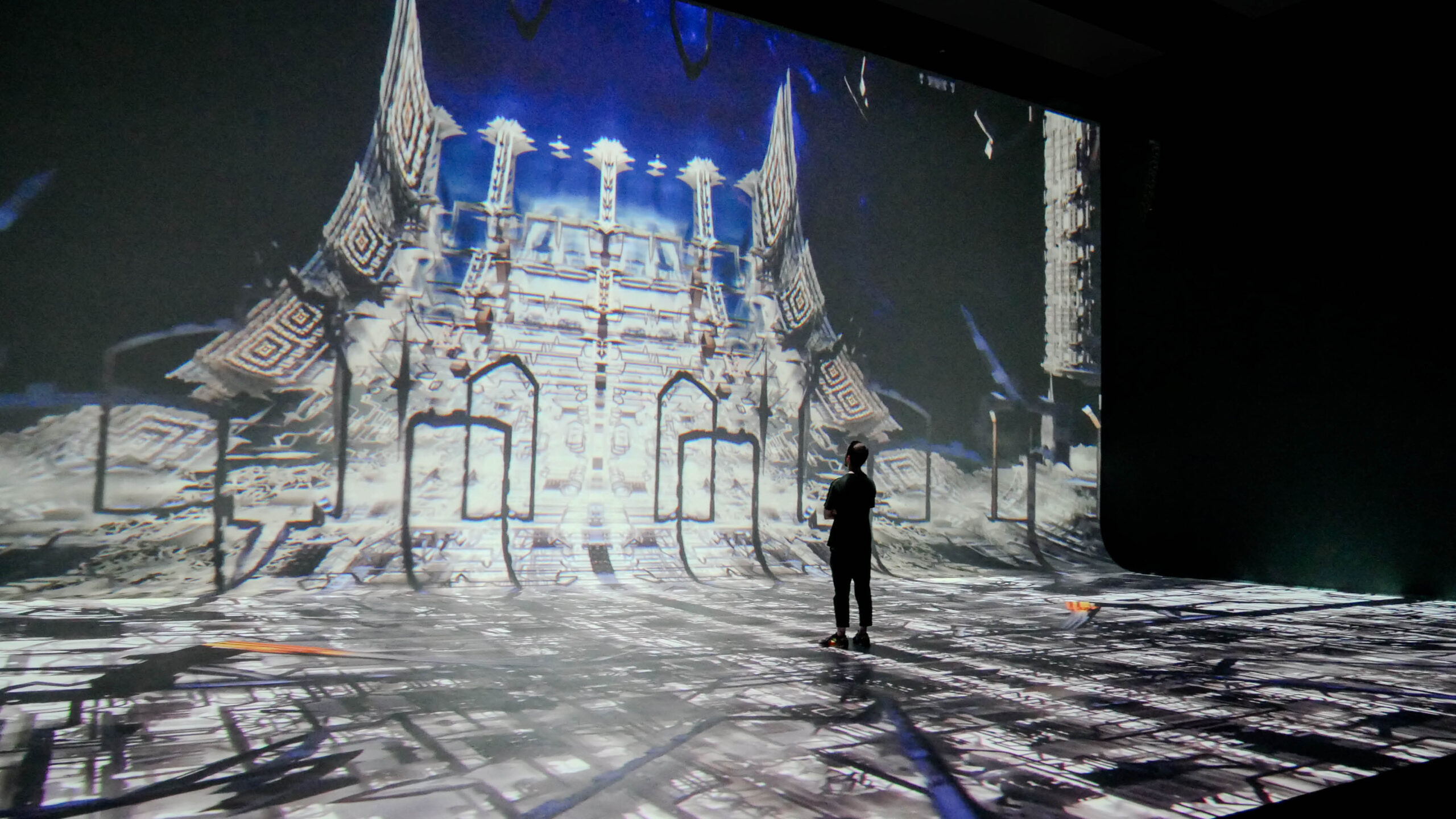 Person silhouetted against large digital projection of intricate, illuminated architectural structure in dark exhibition space