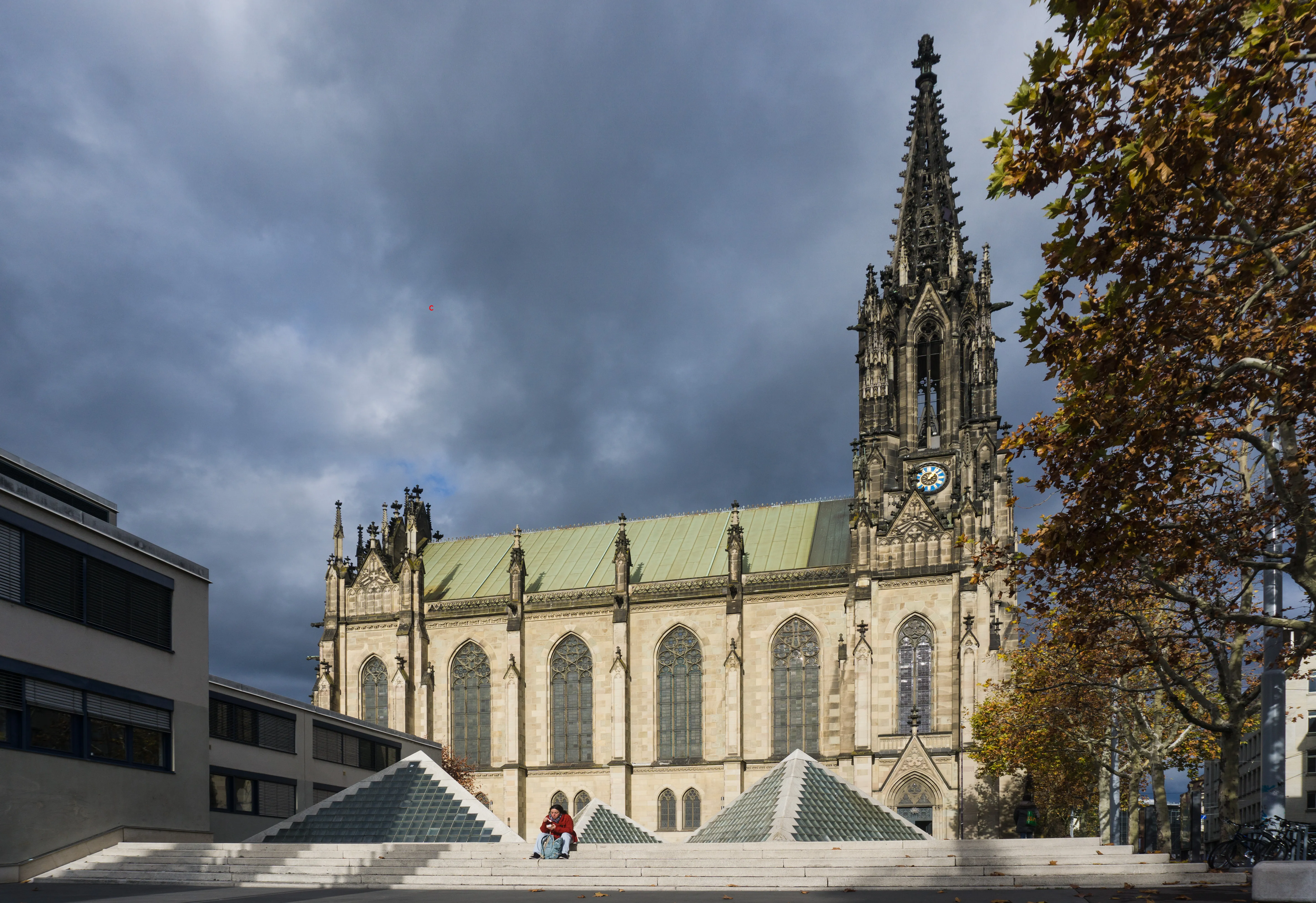 Buitenaanzicht van de neogotische Elisabethenkirche met zijn hoge torenspits en groene dak, omlijst door moderne piramidevormige daklichten op de voorgrond onder een bewolkte lucht.