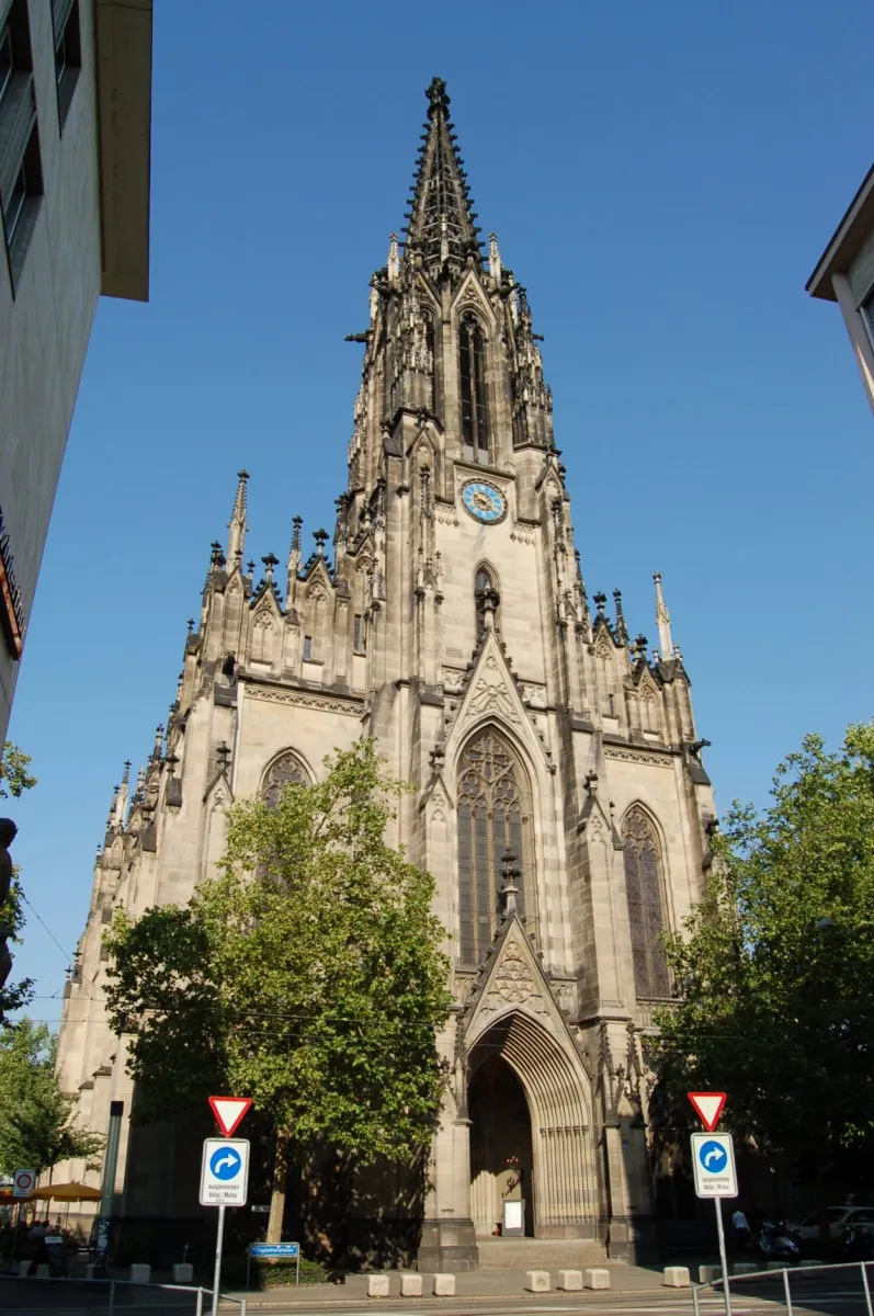 Hoge neogotische kerk met een puntige toren, een wijzerplaat en een boogvormige ingang omlijst door groene bomen onder een heldere blauwe lucht