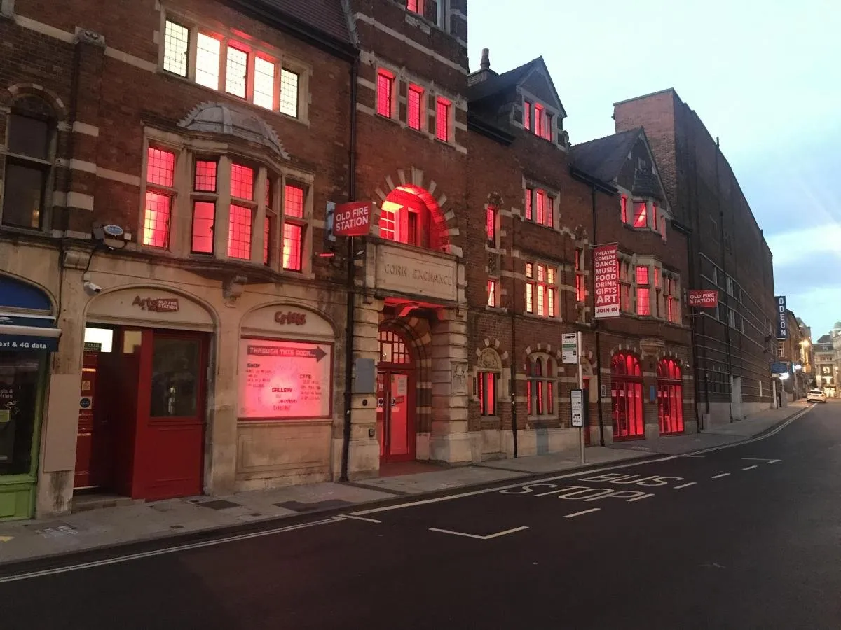 Widok z poziomu ulicy na budynek z czerwonej cegły o zmierzchu, z oknami świecącymi jasną czerwienią; widoczna fasada centrum sztuki Old Fire Station z łukowatym wejściem i oznaczeniami ulicy.
