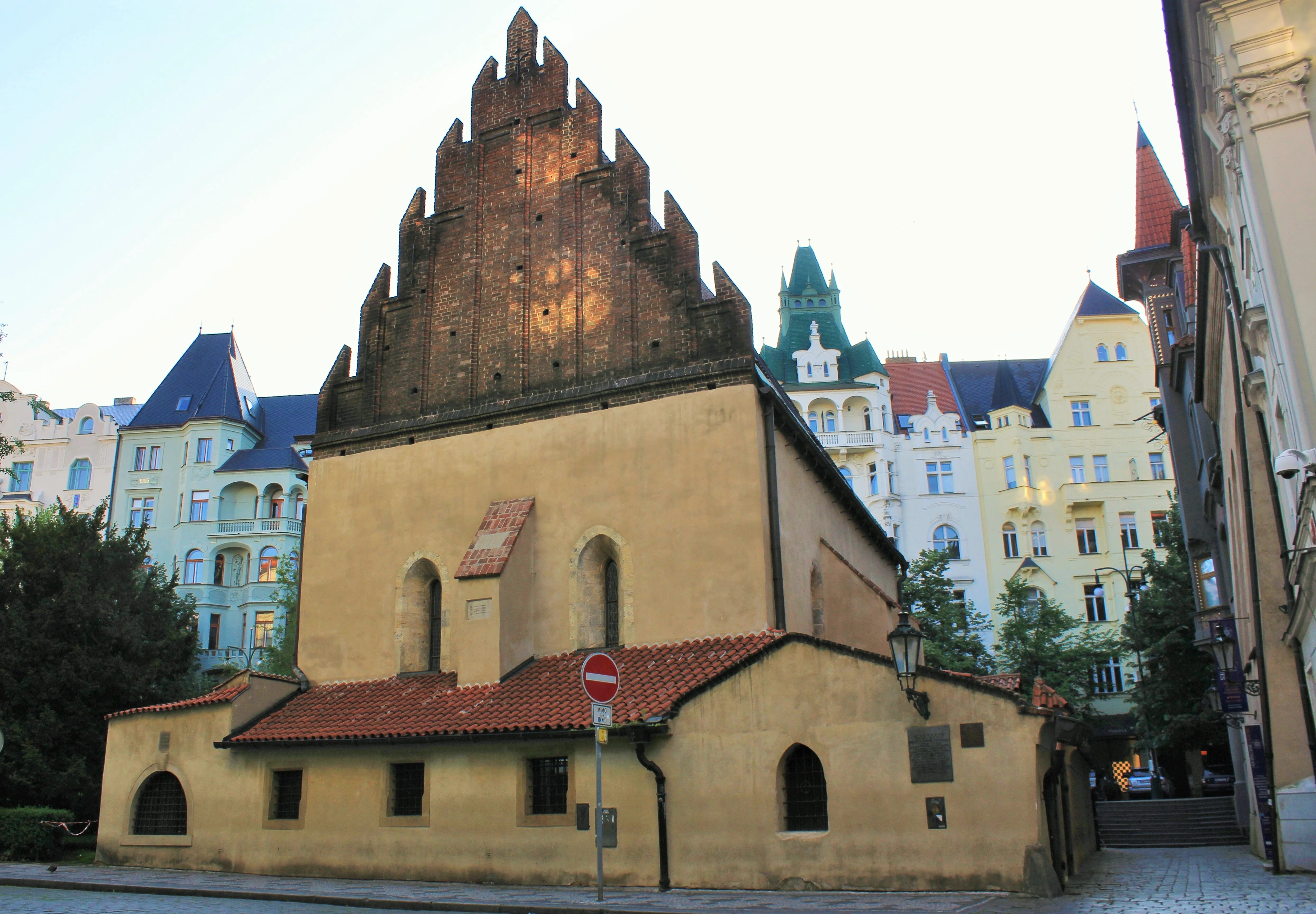 La Sinagoga Vieja-Nueva de Praga con un hastial escalonado y ventanas arqueadas, rodeada de edificios coloridos.