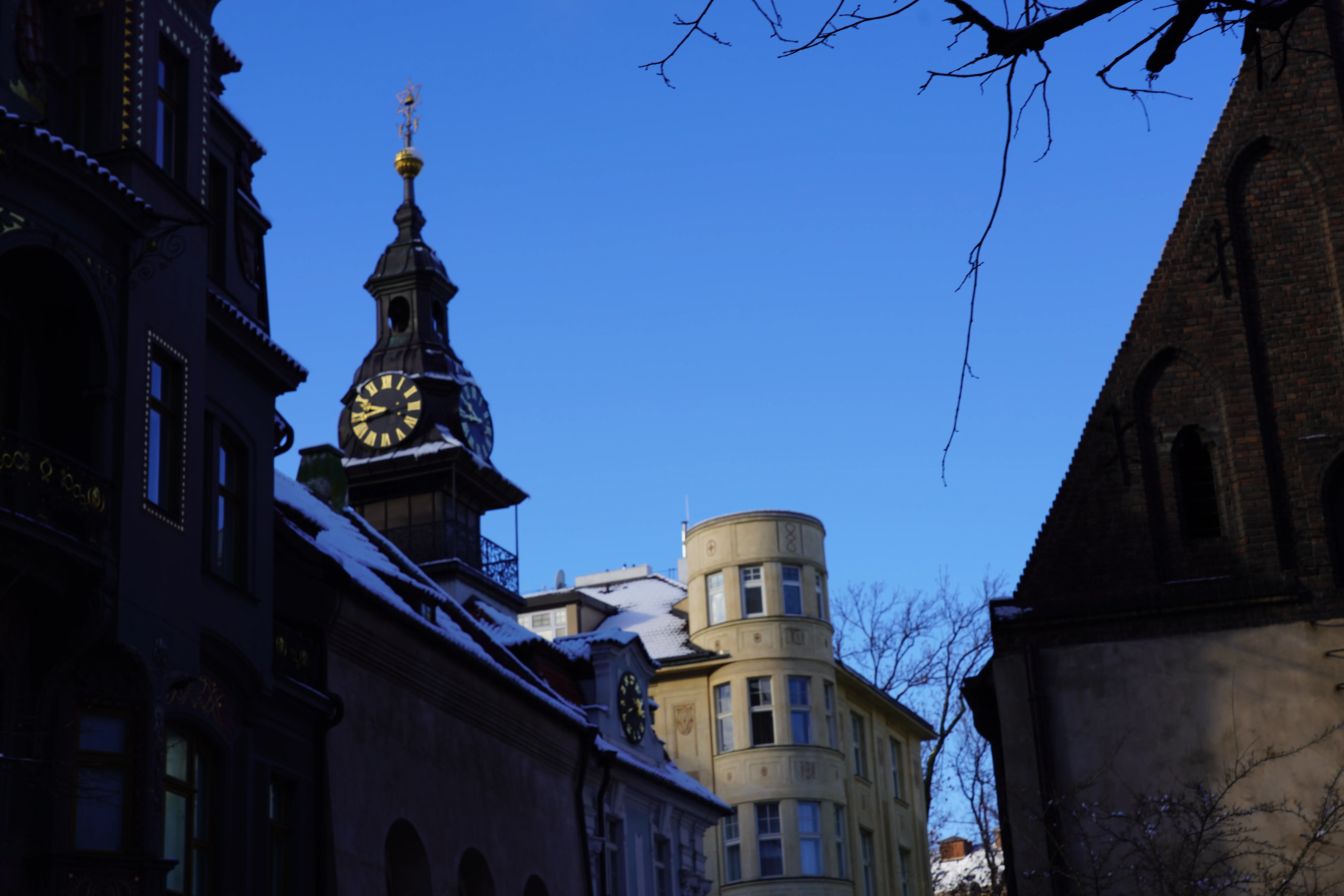 Een smalle straatweergave met de toren van de Oude-Nieuwe Synagoge met een klok, omringd door historische gebouwen onder een helderblauwe lucht.