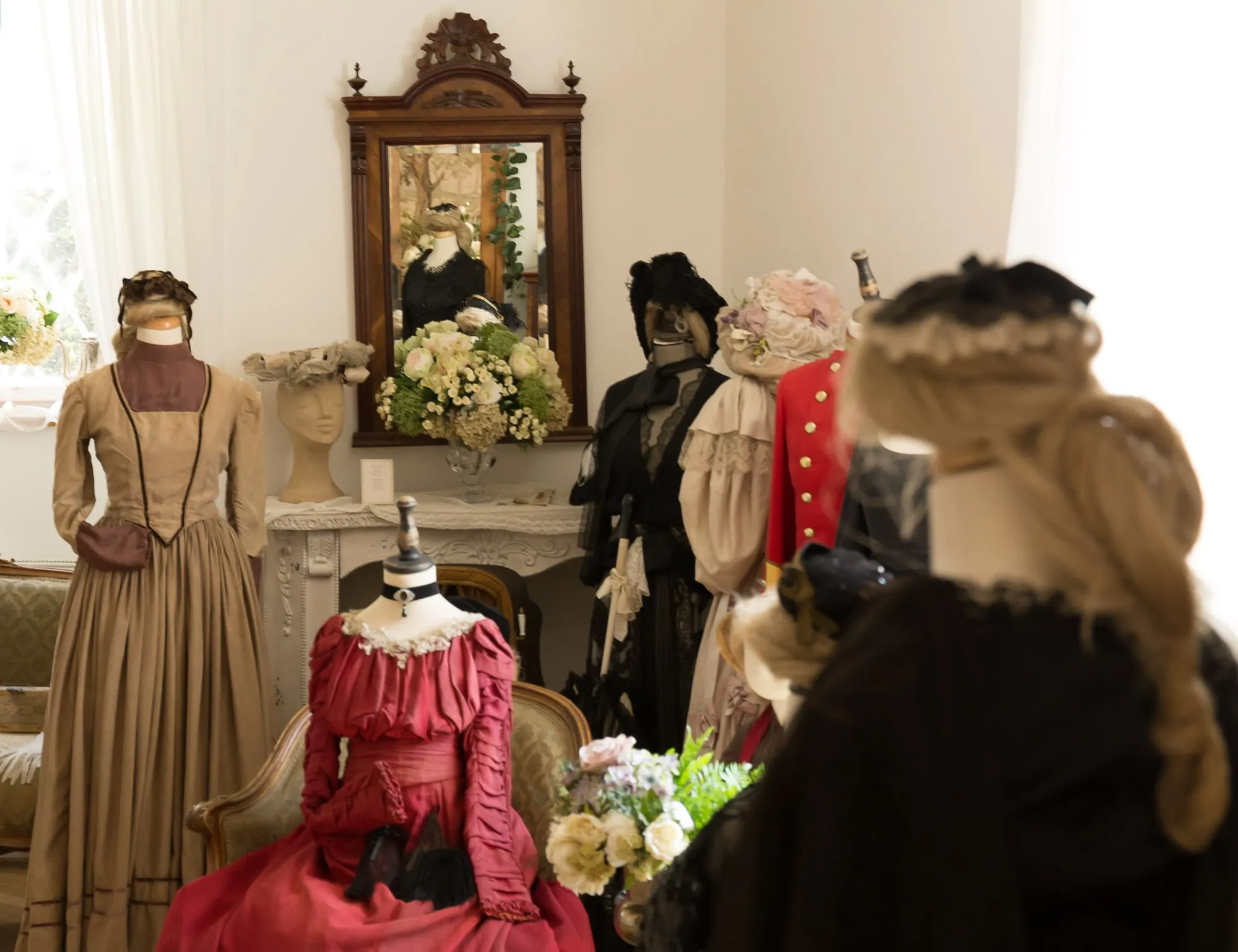 Maniquíes con trajes históricos, que incluyen un vestido beige y un vestido rojo, expuestos en una sala de museo con un espejo y flores