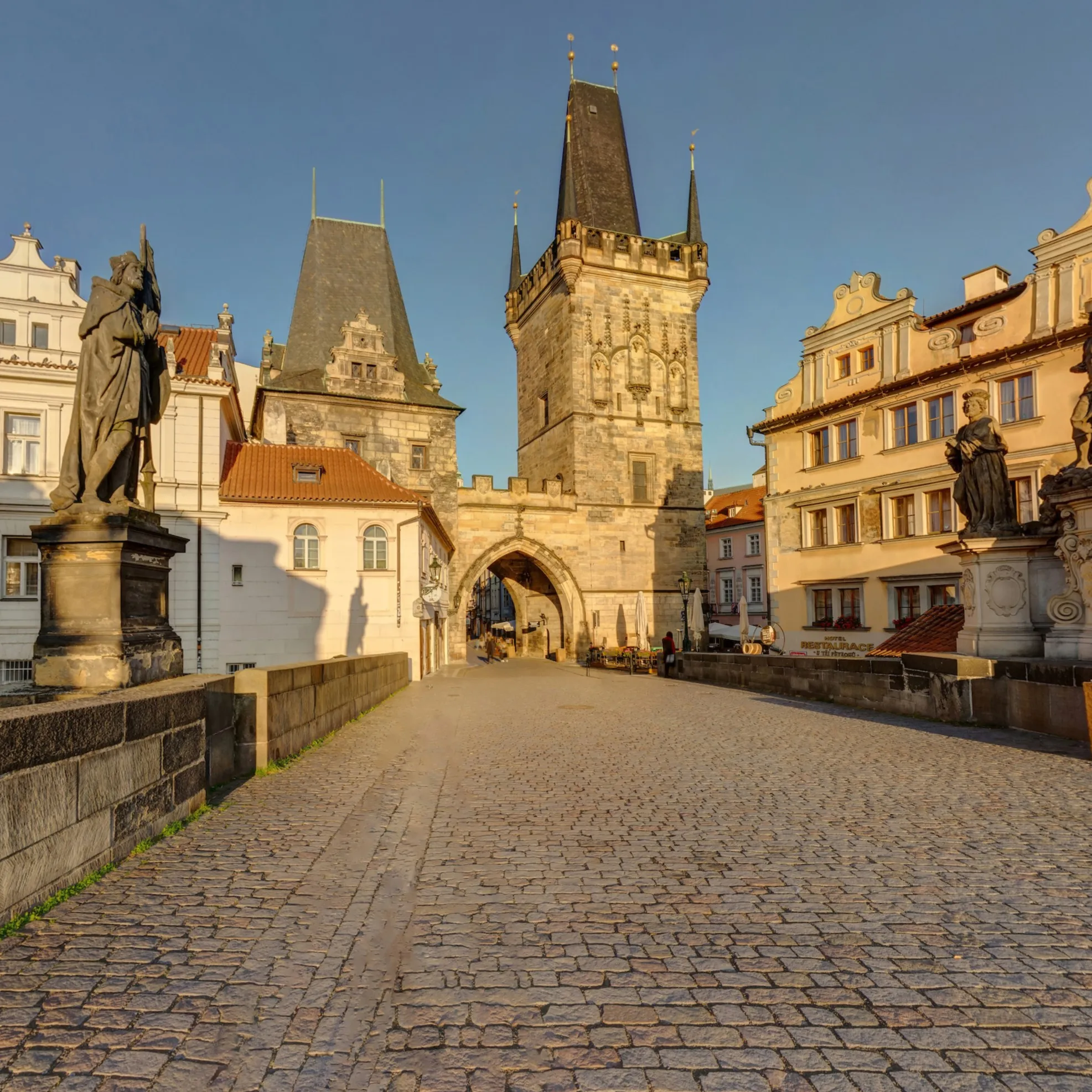 Gothic stone tower with spires at the end of a cobblestone bridge, flanked by statues and historic buildings under a clear sky