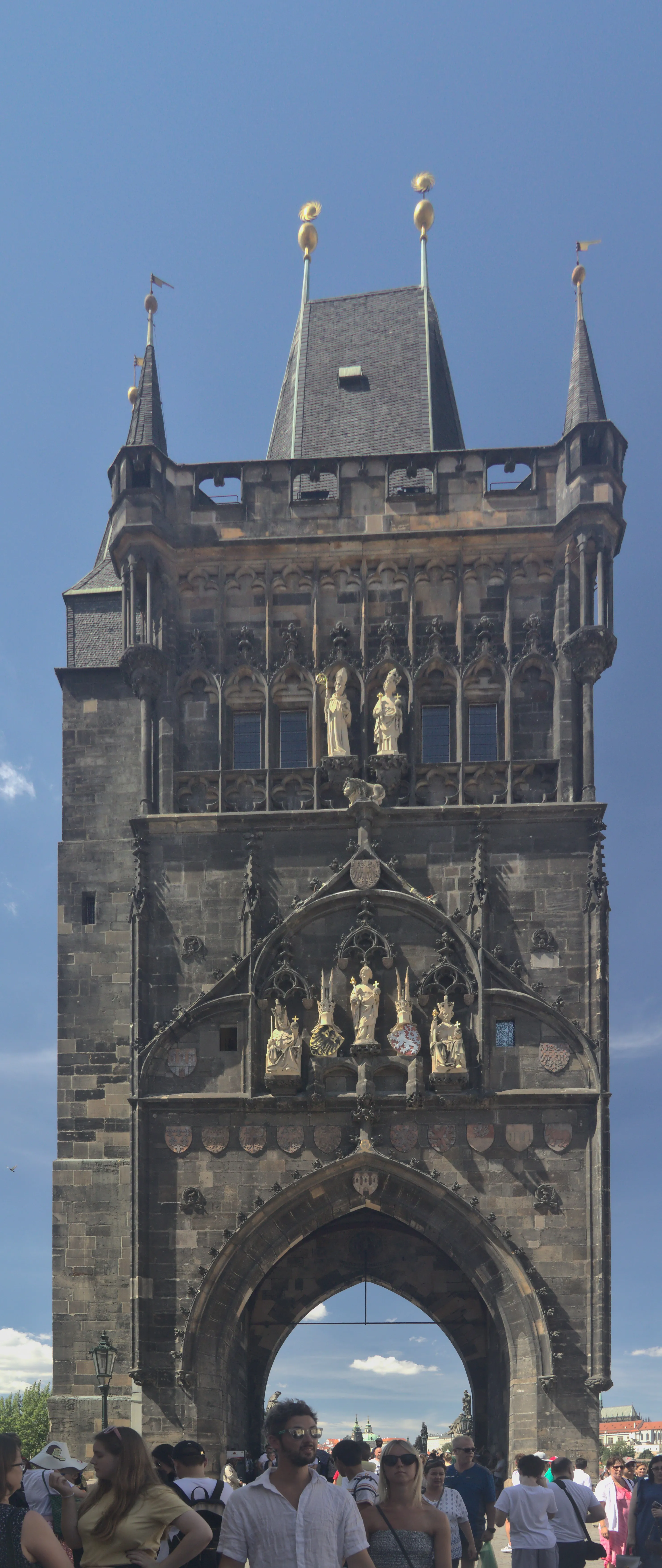 Gothic stone tower with spires and statues under a blue sky, people at the base