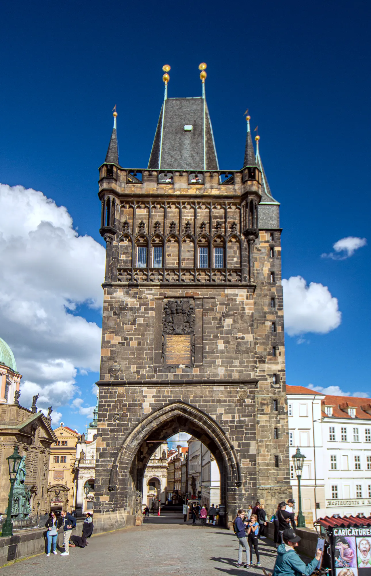 A tall Gothic stone tower with a large arched entrance, spires, and golden finials against a blue sky with white clouds.
