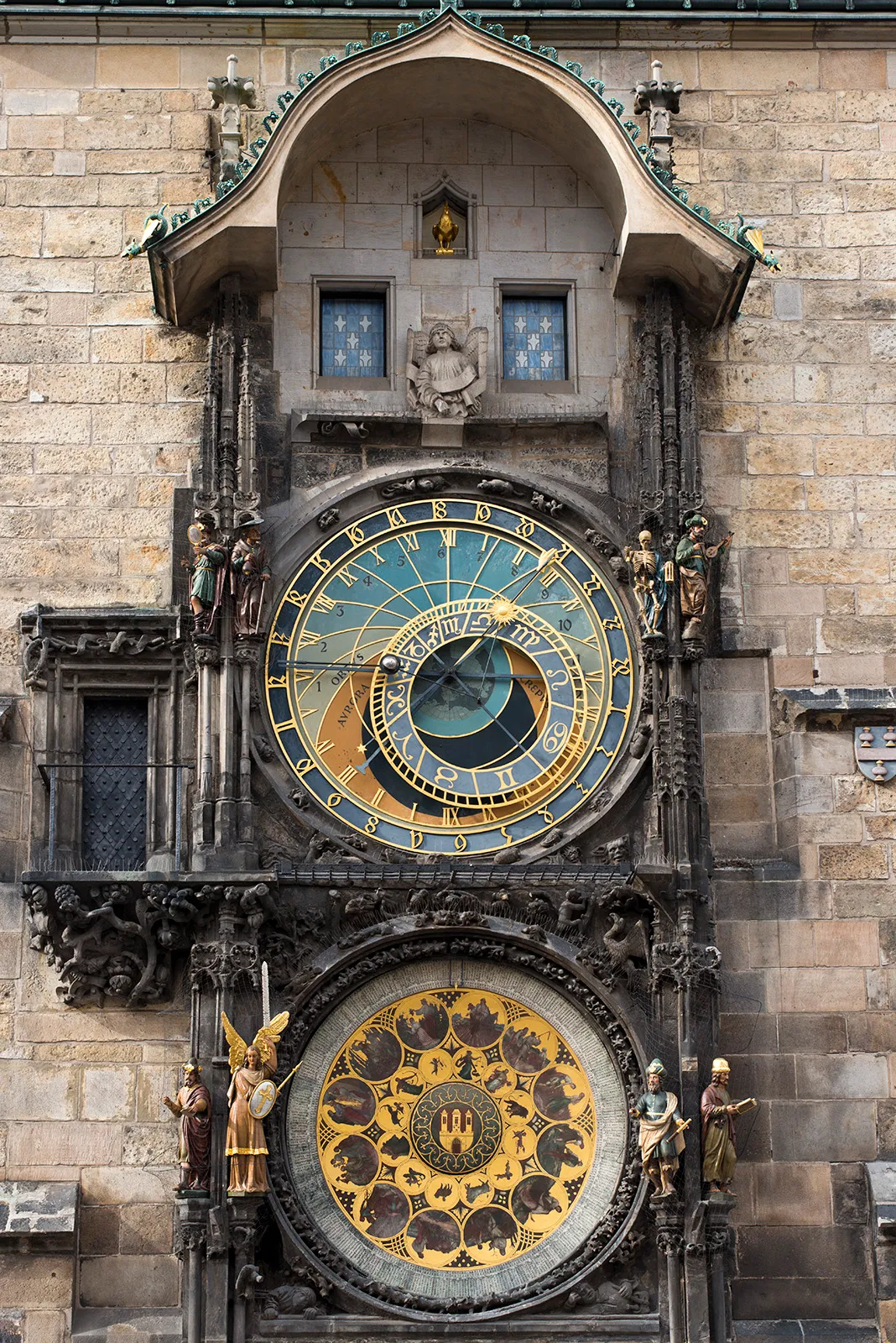 Zbliżenie na praski zegar astronomiczny na Ratuszu Staromiejskim, ukazujące jego ozdobny projekt ze złotymi figurami, niebieskimi i złotymi tarczami oraz kamienną architekturą.