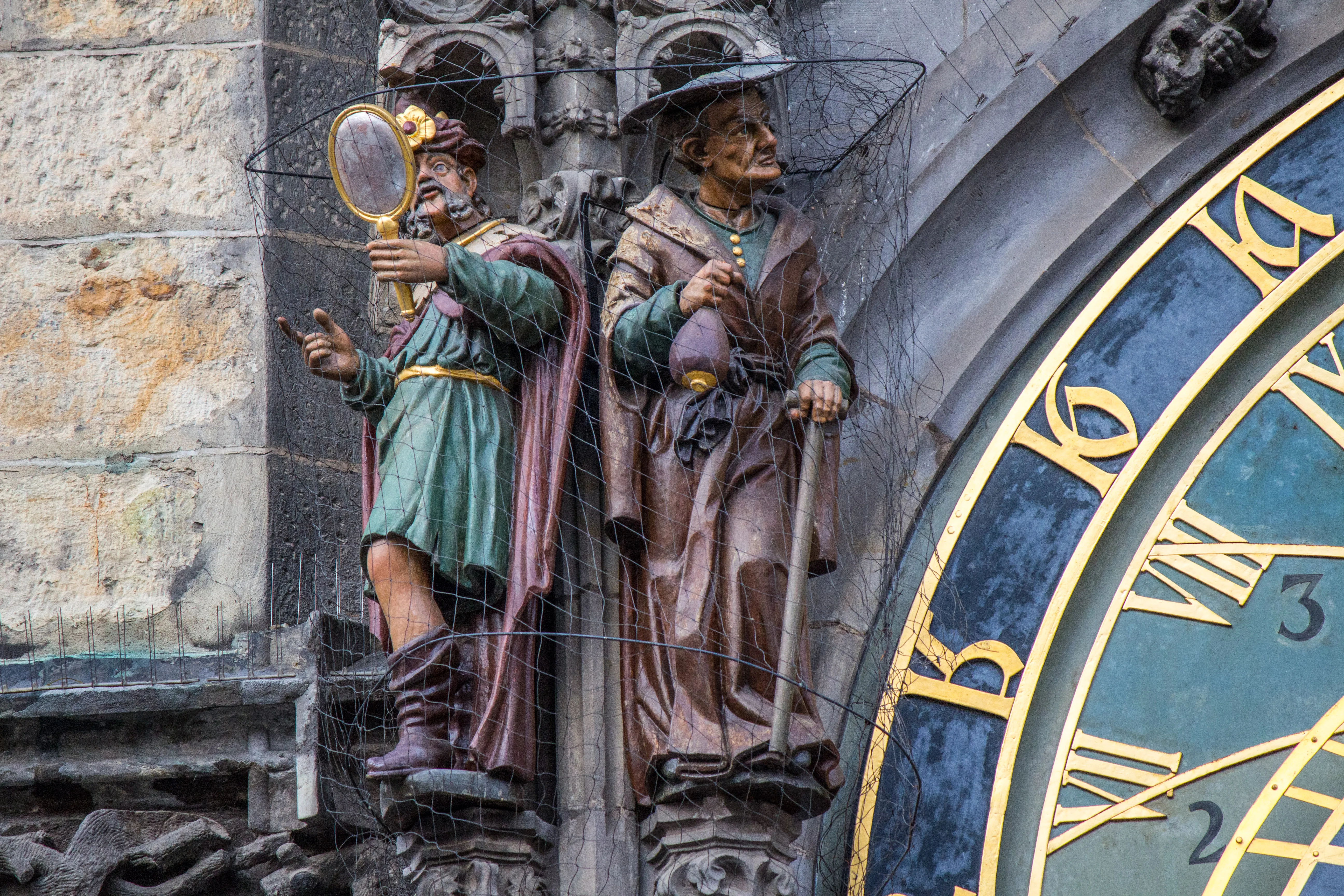 Zbliżenie dwóch ozdobnych posągów na zegarze astronomicznym w Pradze, jeden trzymający lustro, a drugi latarnię, obok niebieskiej tarczy zegara ze złotymi rzymskimi cyframi.