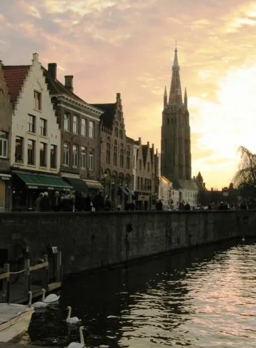 Escena del canal en Brujas al atardecer, con edificios históricos de ladrillo a lo largo del muelle y la alta aguja gótica de la Iglesia de Nuestra Señora al fondo, con cisnes flotando en el agua