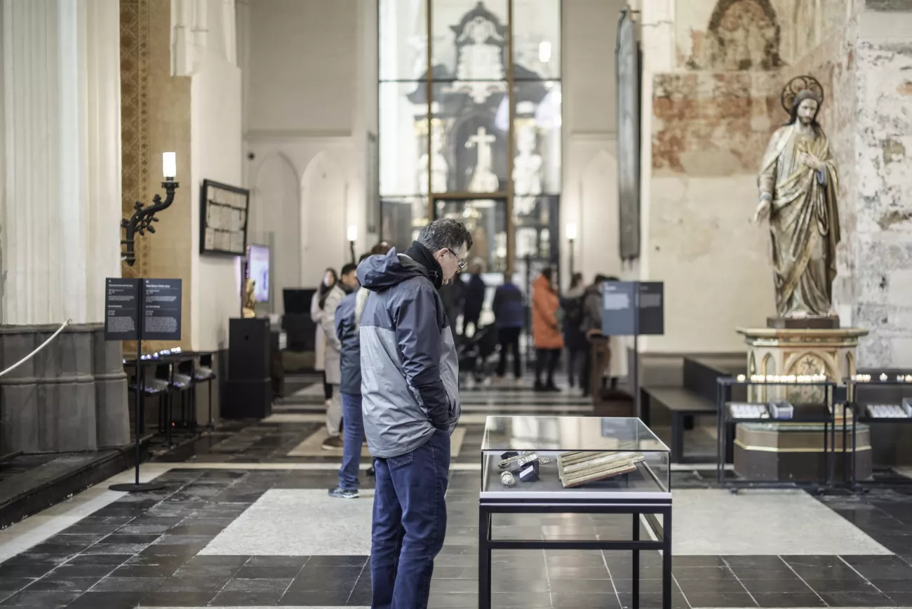Zwiedzający poznają wnętrze Museum Onze-Lieve-Vrouwekerk, w którym znajduje się złocona figura na cokole oraz szklane gabloty wystawowe w nawie o kamiennej posadzce.