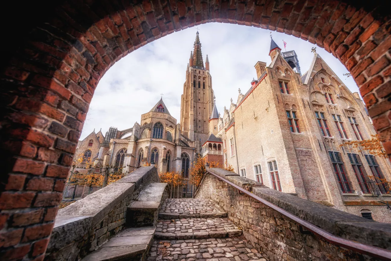 Vista de la alta aguja gótica de la Iglesia de Nuestra Señora y los edificios históricos adyacentes, enmarcada a través de un arco de ladrillo con escalones de adoquines