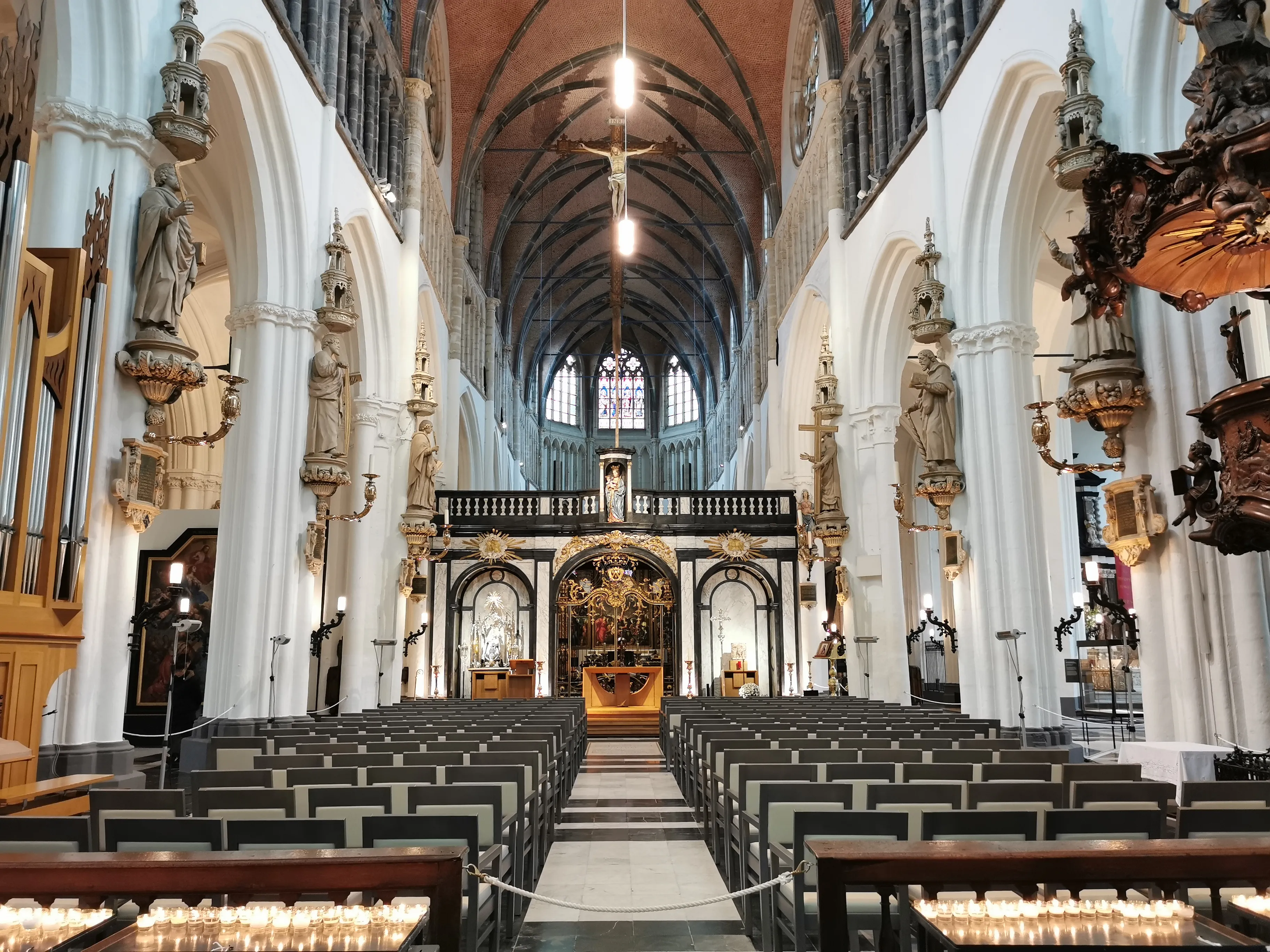 Interior de la Iglesia de Nuestra Señora en Brujas, que muestra la nave con hileras de bancos, columnas blancas con estatuas y el altar ornamentado.