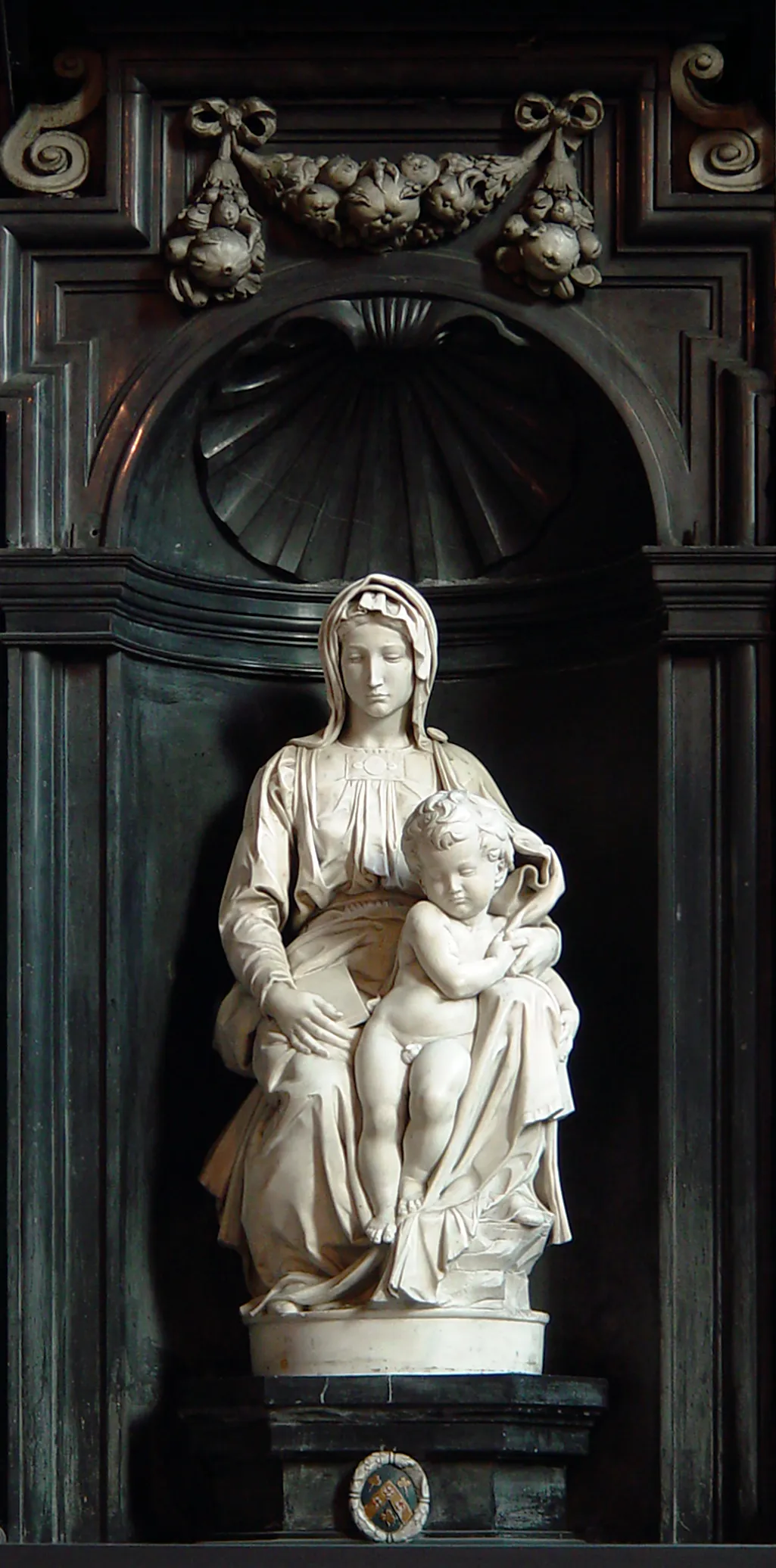 Escultura de mármol blanco de la Virgen María sentada con el Niño Jesús, situada dentro de una hornacina arquitectónica oscura y ornamentada con tallas decorativas y un motivo de concha.