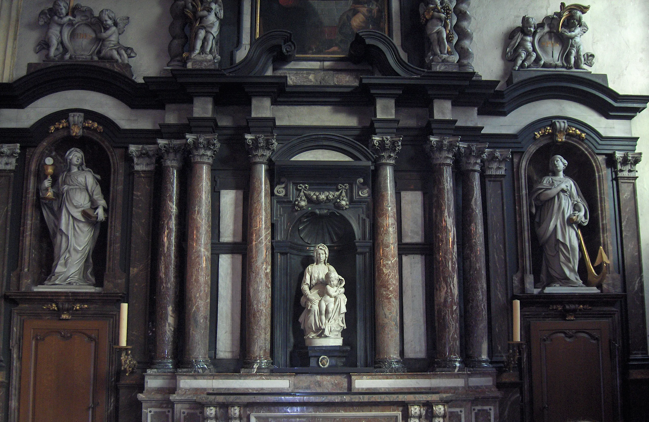 Escultura de mármol blanco de la Virgen con el Niño, obra de Miguel Ángel, situada en el centro de un oscuro y ornamentado altar de iglesia con columnas y estatuas a los lados en hornacinas