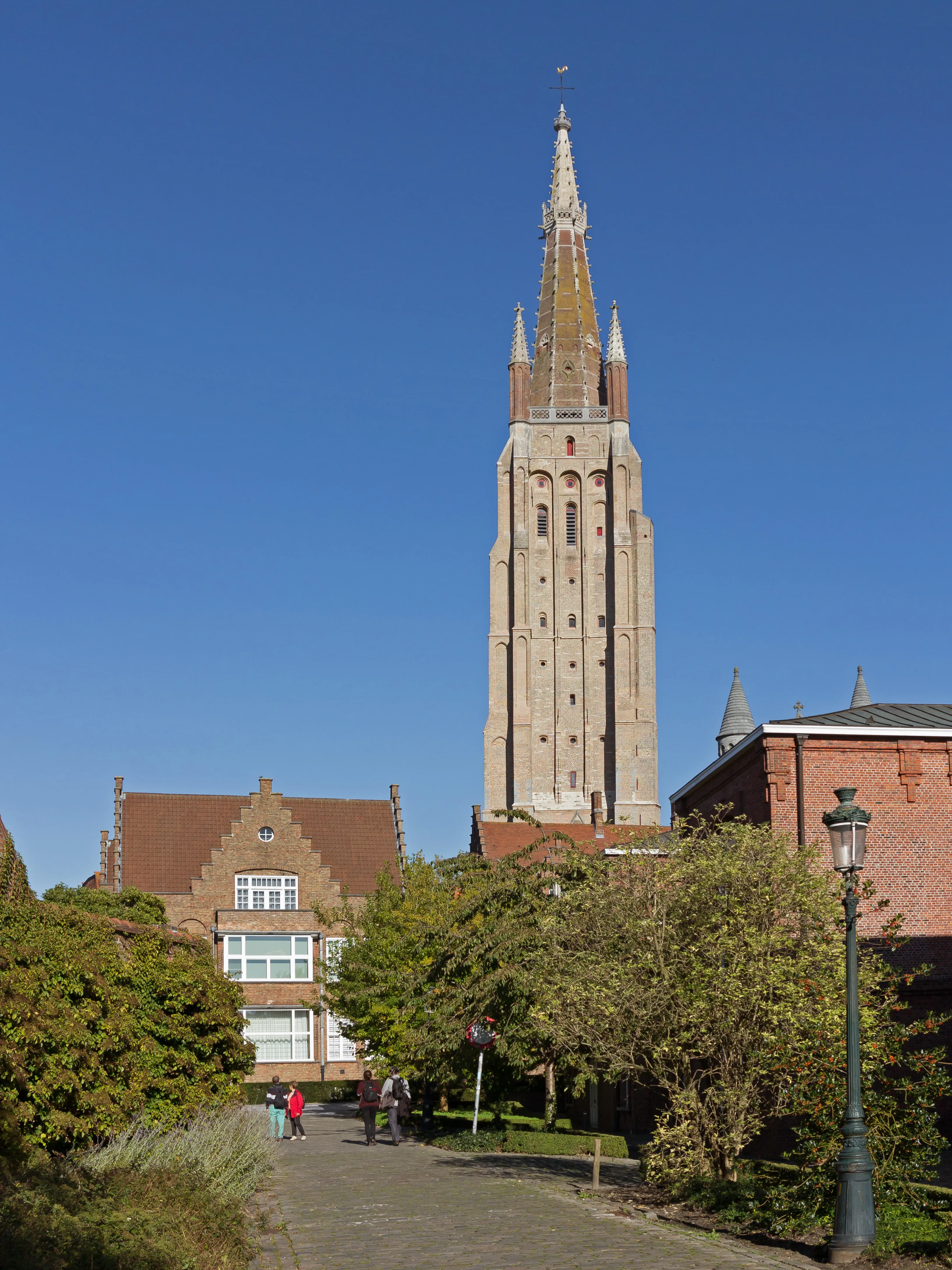 Alta torre de iglesia de ladrillo con una aguja ornamentada que se alza sobre los edificios y árboles circundantes bajo un cielo azul despejado