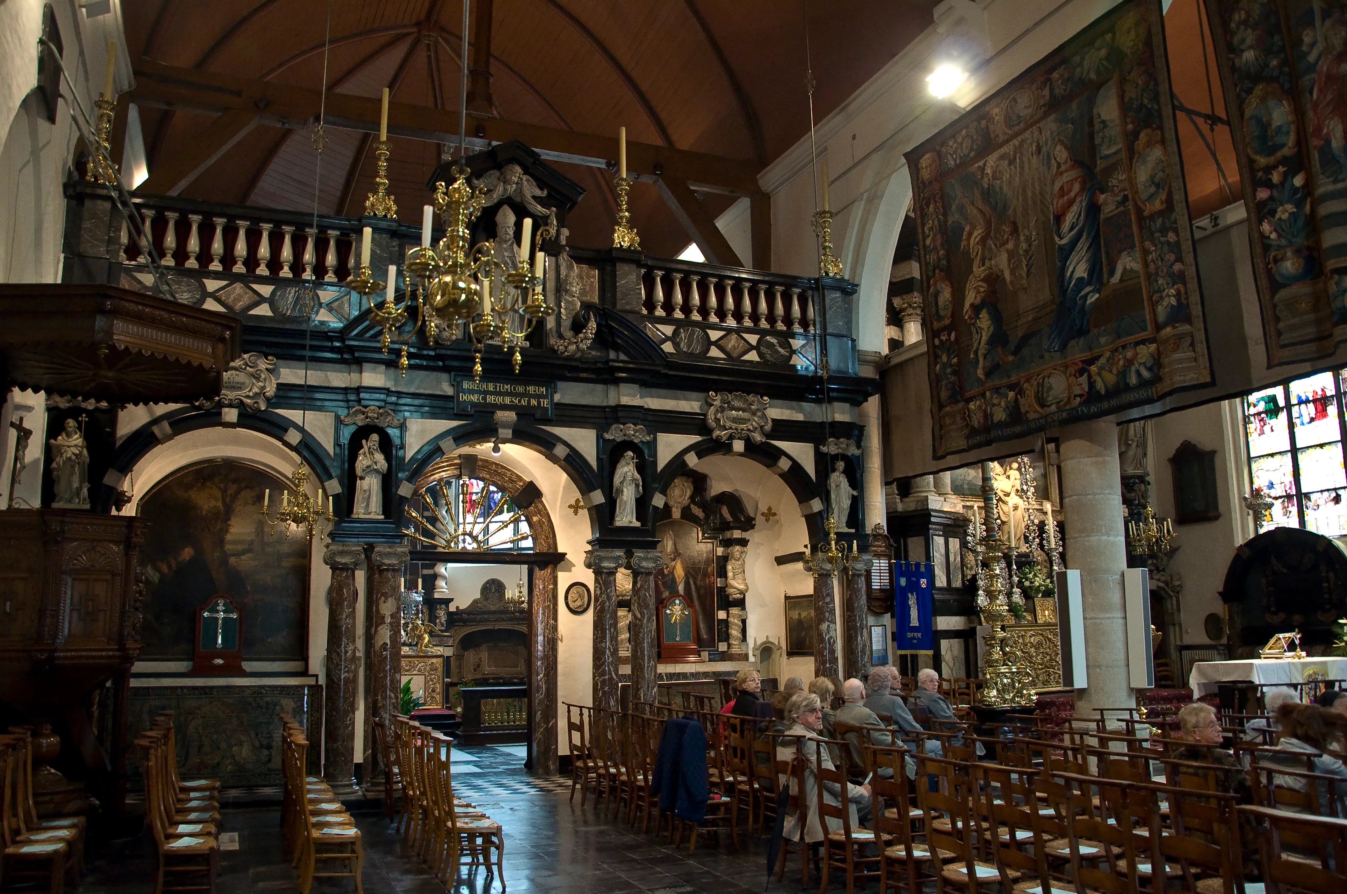 Interior de la iglesia de Onze-Lieve-Vrouw ter Potterie que muestra un ornamentado cancel de madera con estatuas, una gran lámpara de araña, bancos de madera y tapices en las paredes.