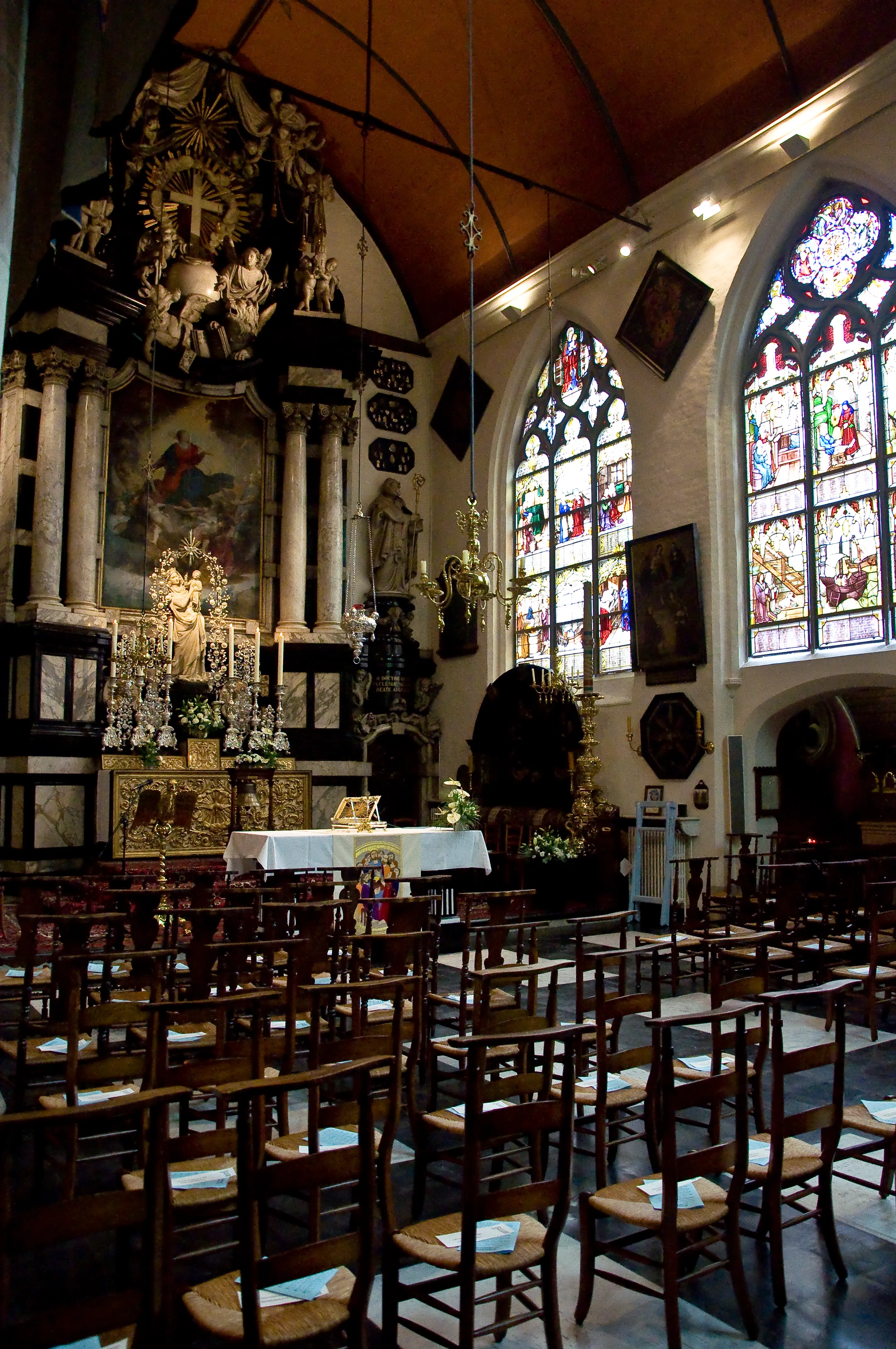 Interior de la iglesia de O.L.V. ter Potterie que muestra un ornamentado altar barroco, filas de sillas de madera y altos vitrales.
