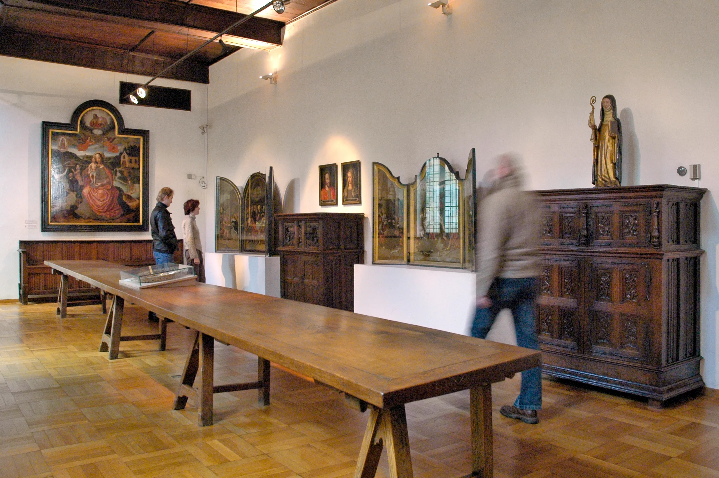 Interior de una sala de museo con una larga mesa de caballete de madera, aparadores de madera tallada a lo largo de las paredes, pinturas de retablos religiosos y una estatua dorada, con visitantes observando las exposiciones