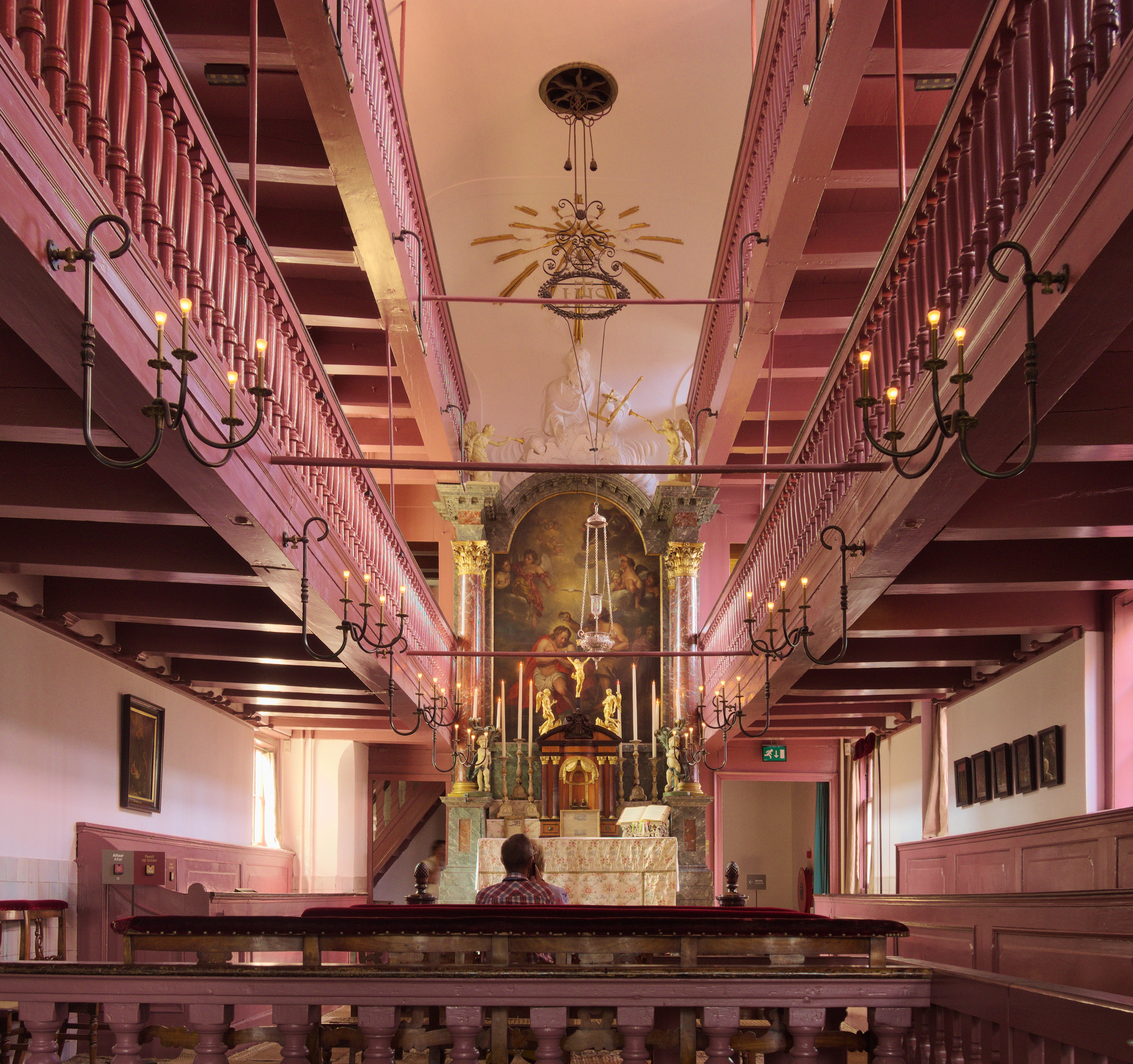 Interior of a hidden church with pink walls, ornate altar, and hanging chandeliers in a canal house museum.
