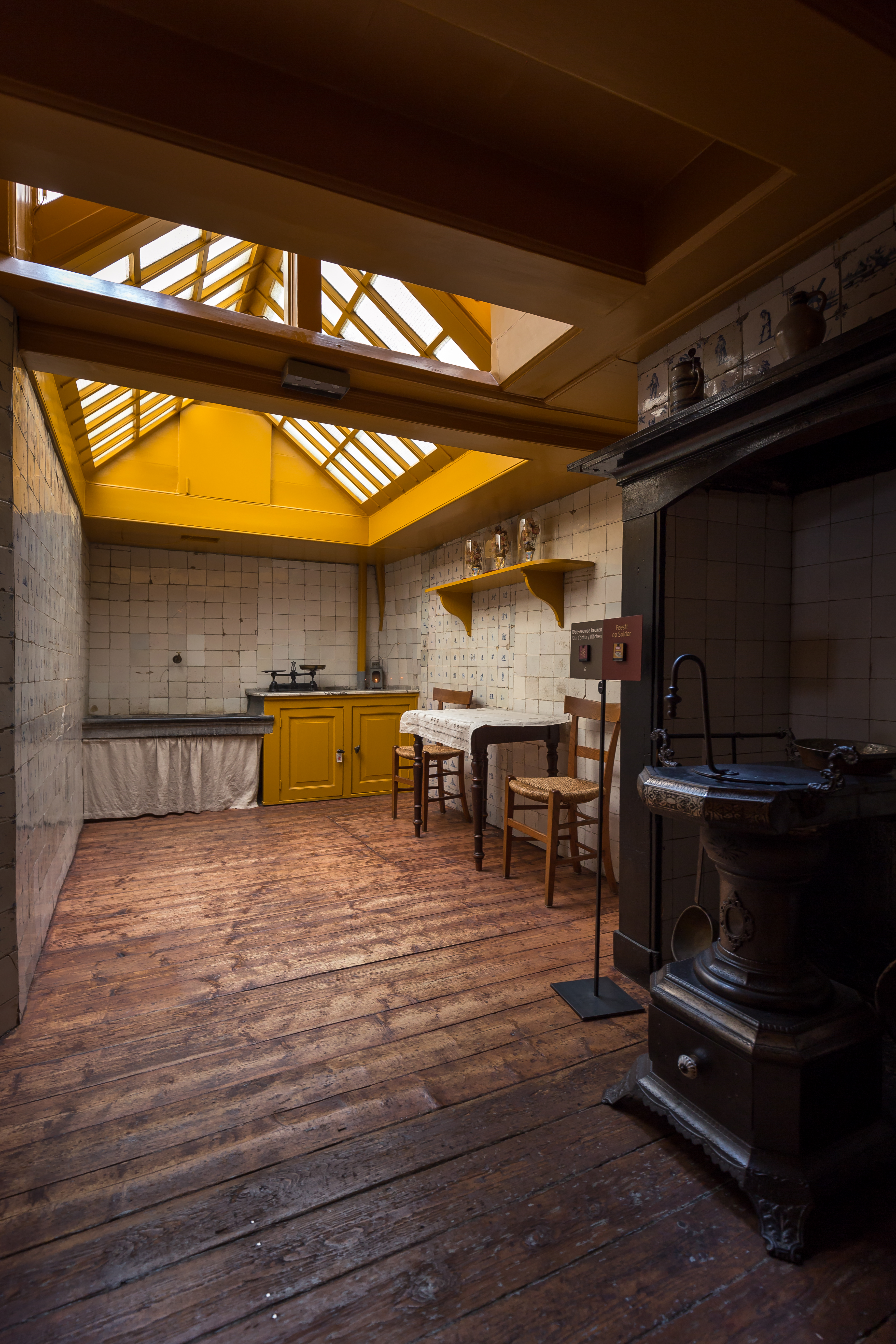Historical kitchen interior with wooden floors, yellow cabinets, dining table, chairs, and large cast-iron stove under skylights