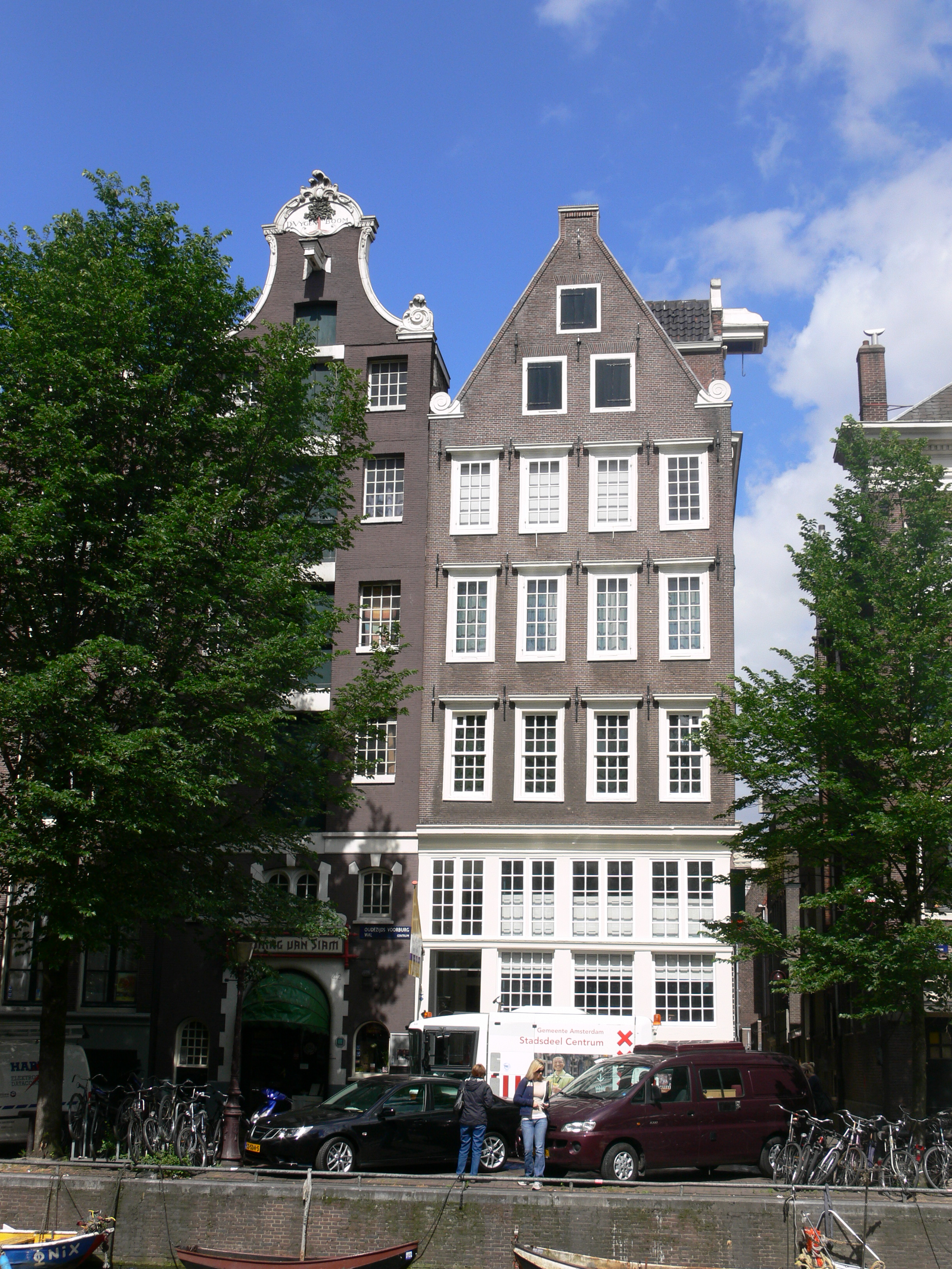 A tall brown canal house with white windows and a stepped gable, situated on a canal with trees and bicycles nearby.