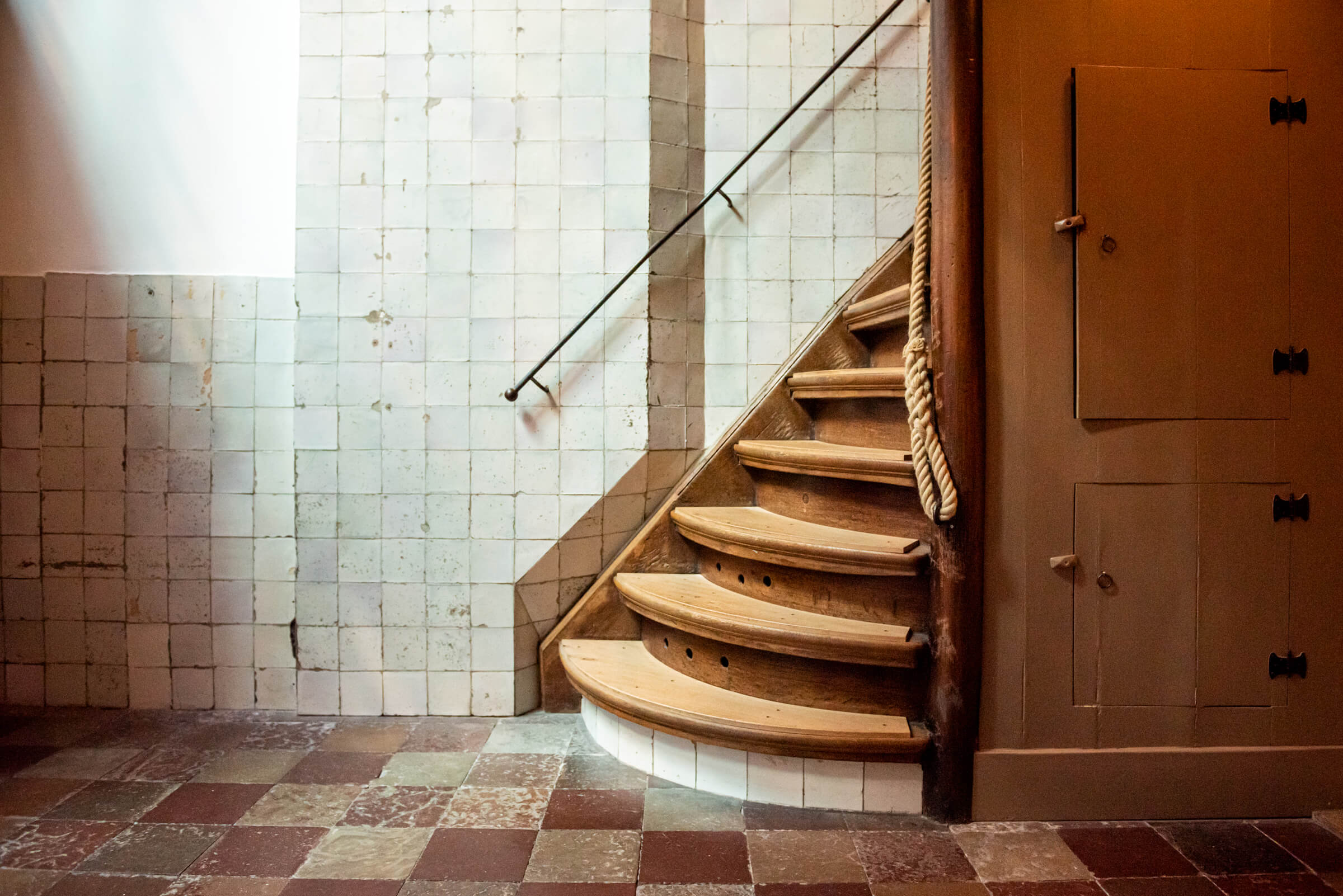 Curved wooden staircase with rope handrail against white tiled walls, leading to wooden door with metal fixtures on checkered tile floor