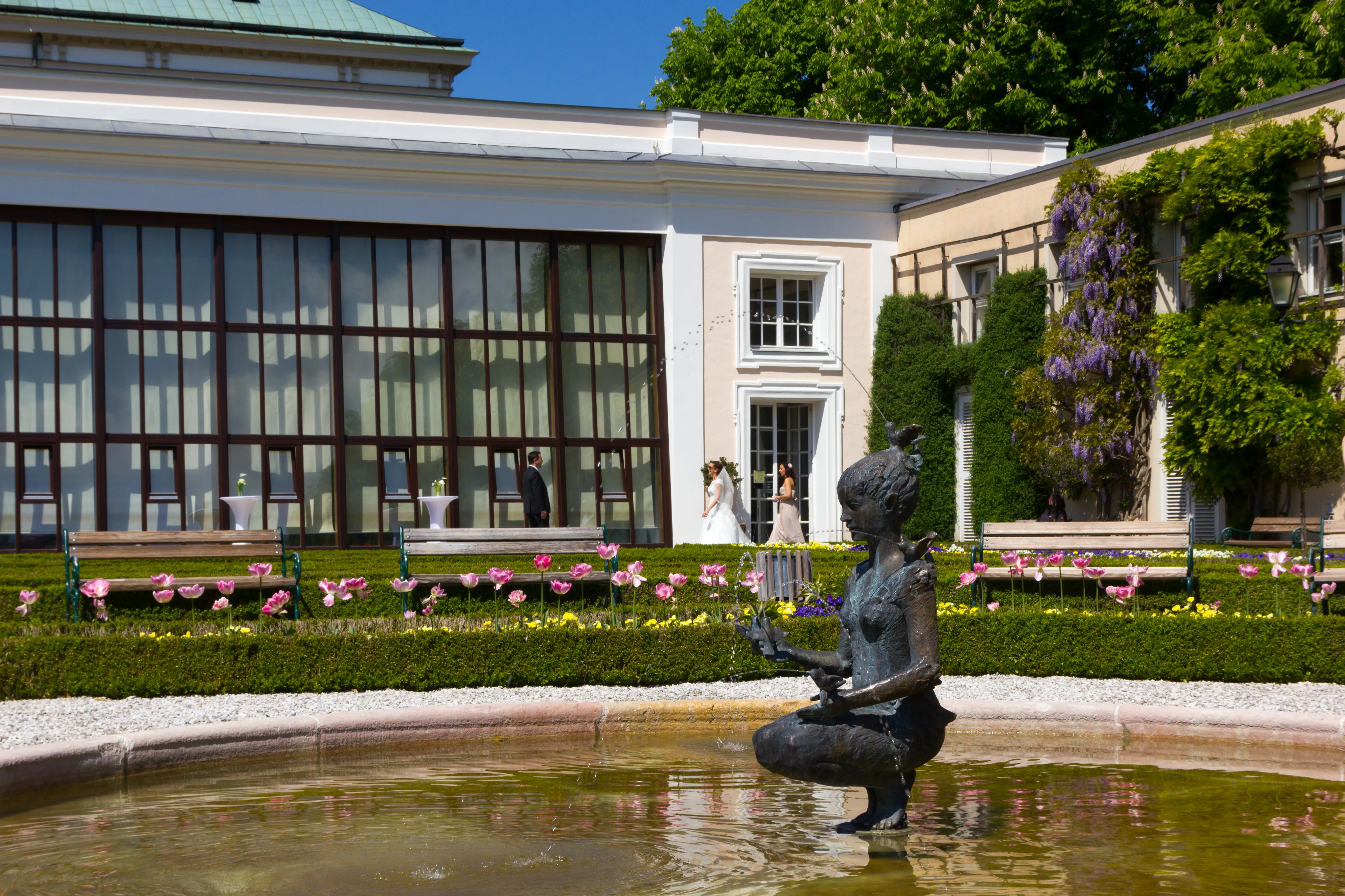 Exterior view of the Orangery Salzburg with large glass windows, a garden fountain featuring a bronze statue, and colorful flower beds