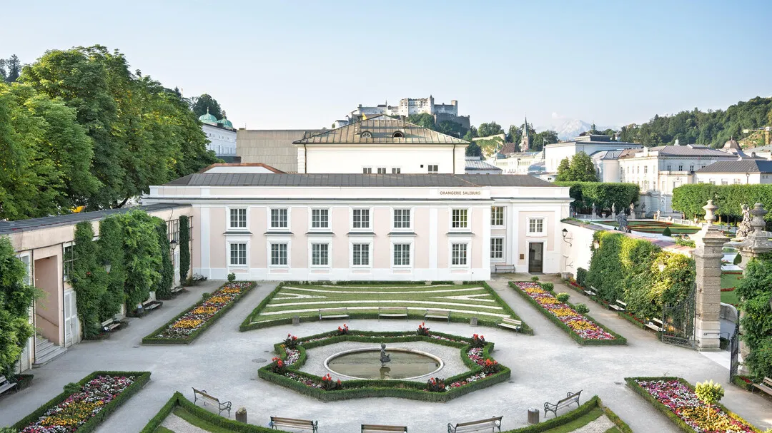 Wide aerial view of the white Orangery building in Mirabell Gardens with formal flower beds, trimmed hedges, a central fountain, and Hohensalzburg Fortress visible on the hill in the background.
