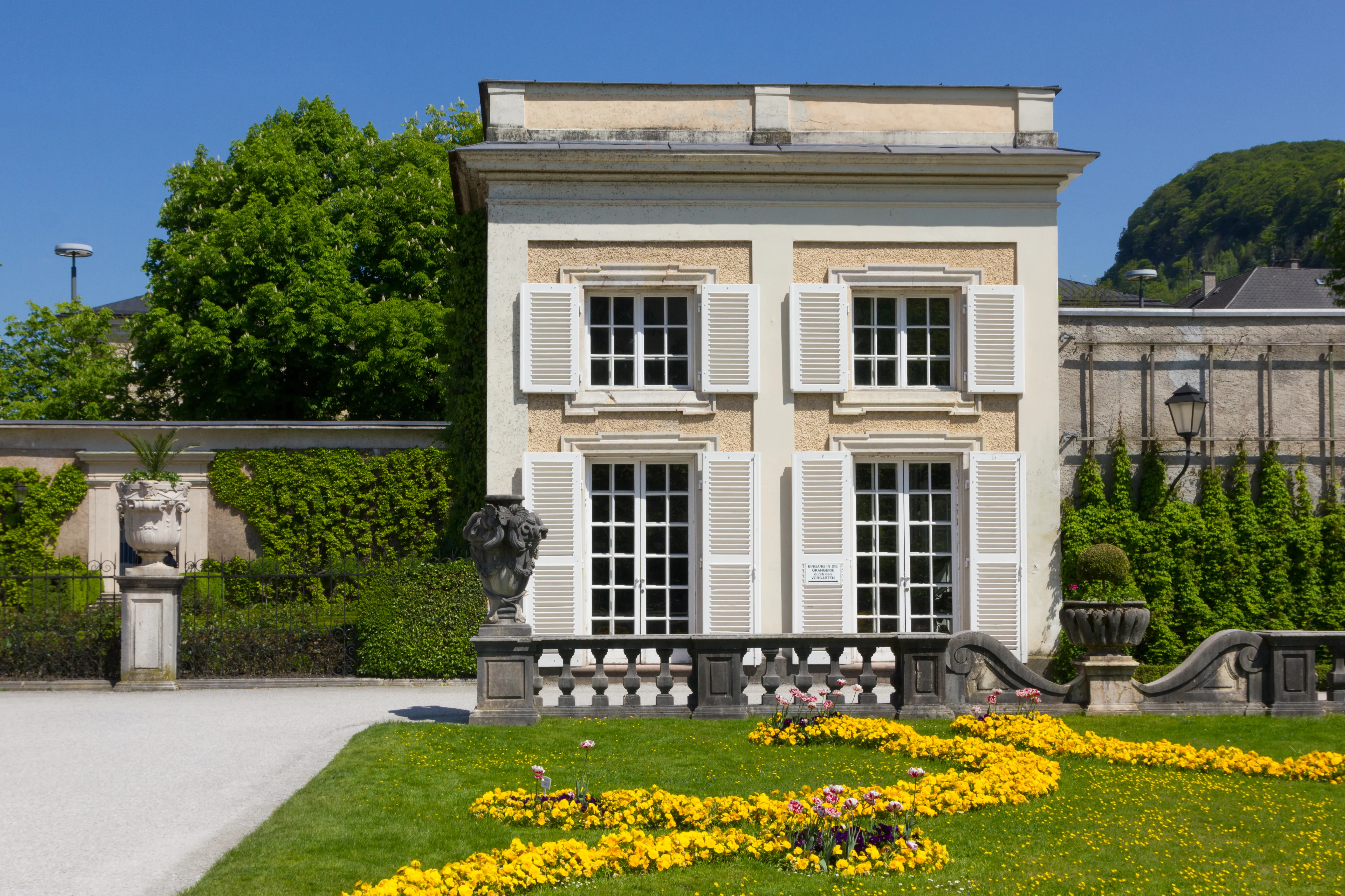 White classical building with shuttered windows and a garden of yellow flowers in front, with a hilltop fortress visible in the background