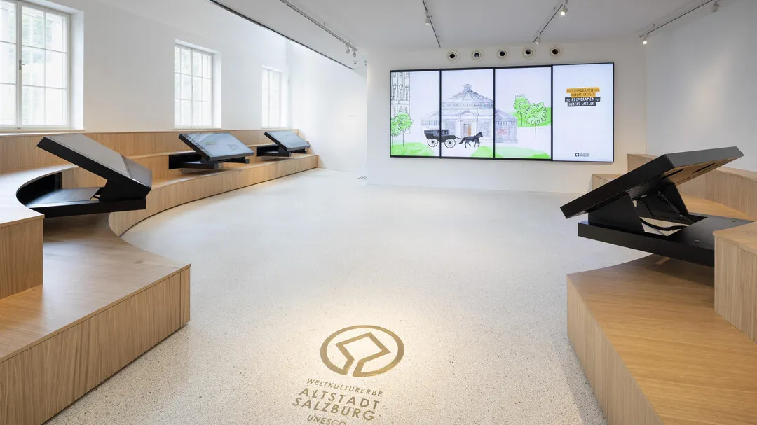 Bright modern exhibition room with curved wooden tiered benches, interactive touch-screen displays, a large multi-panel screen showing a panoramic illustration, and an Altstadt Salzburg logo projected onto the floor.