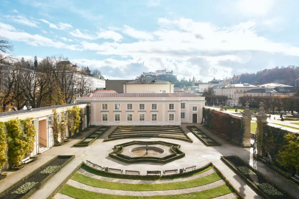 Wide view of the Orangery Salzburg building with its symmetrical formal garden in the foreground and the Hohensalzburg Fortress on the hill in the distance under a blue sky with clouds.