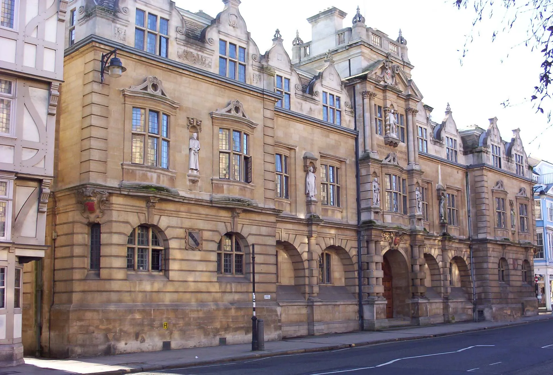 Widok z poziomu ulicy na Rhodes Building przy Oriel College, duży, ozdobny kamienny budynek z łukowatymi oknami na parterze, prostokątnymi oknami na wyższych piętrach, dekoracyjnymi szczytami i posągami we wnękach.