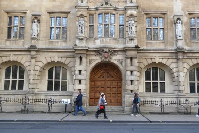 Kamienna fasada Oriel College z łukowymi oknami, posągami w niszach i dużymi drewnianymi drzwiami wejściowymi, z pieszymi idącymi po chodniku