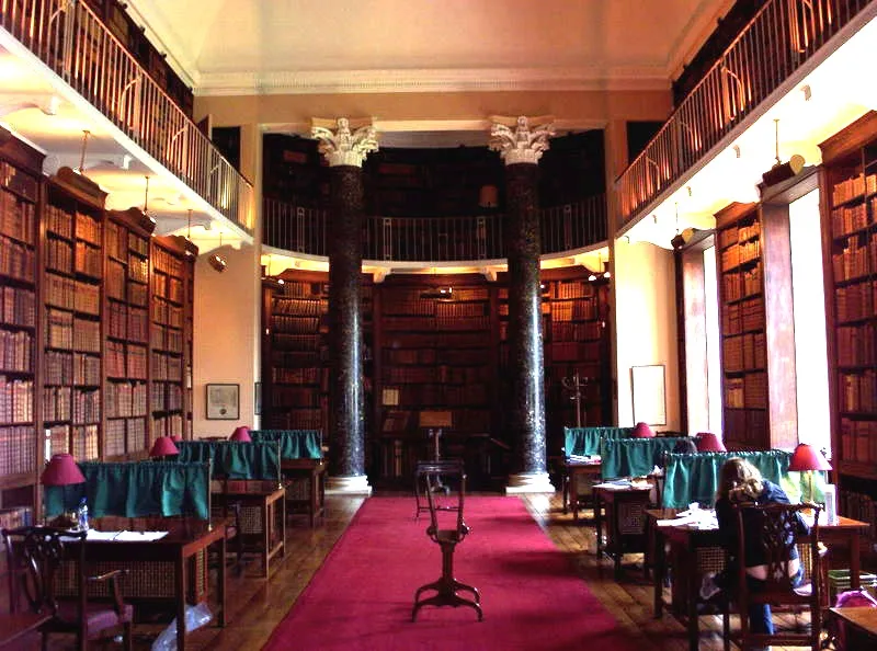 Wnętrze zabytkowej biblioteki z regałami sięgającymi od podłogi do sufitu, dwiema dużymi ciemnymi kolumnami, pulpitami do czytania z zielonymi obrusami i czerwonymi lampami oraz czerwonym chodnikiem biegnącym przez środek.