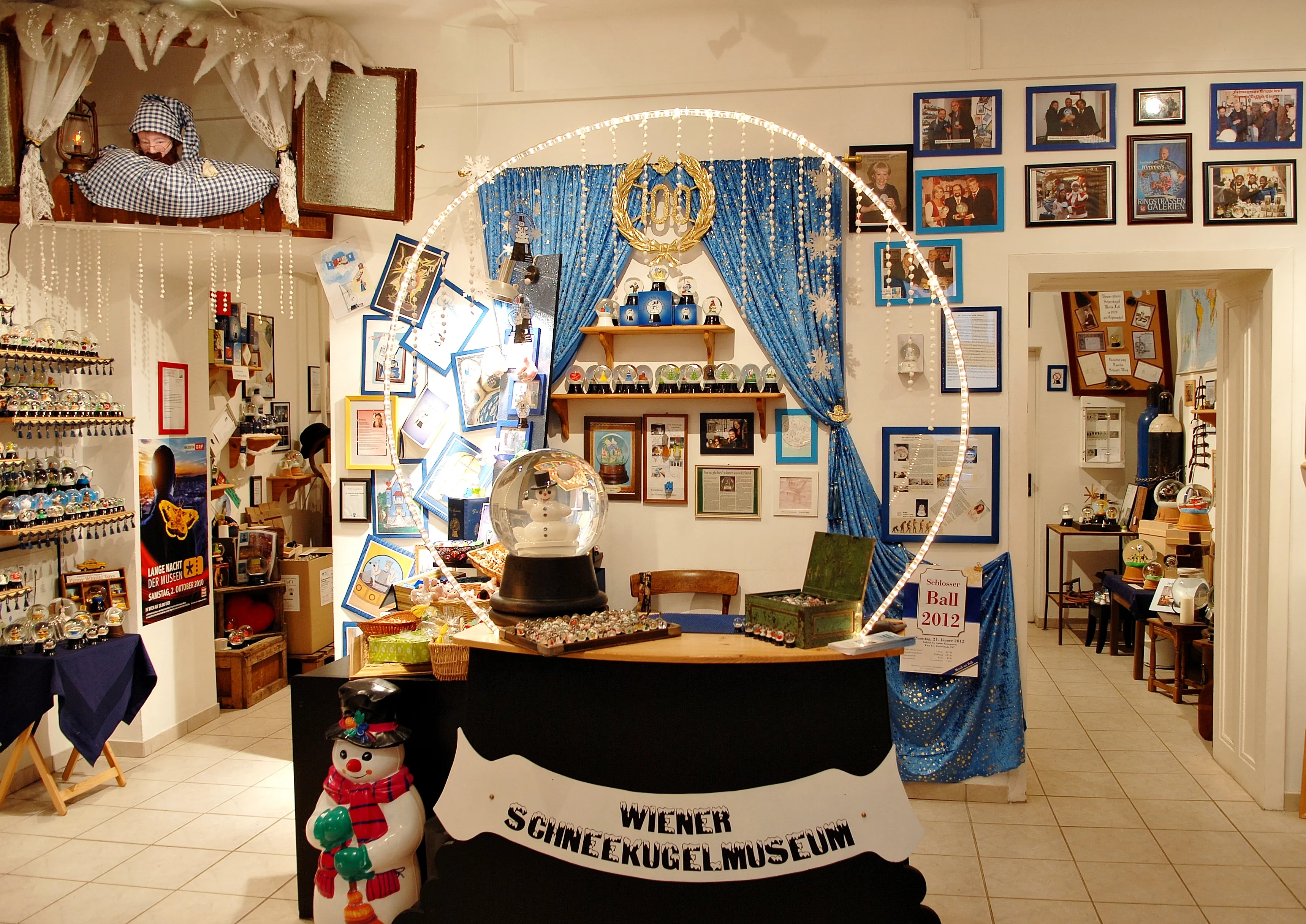 Interior of a museum with a snow globe display, a sign that reads 'WIENER SCHNEEKUGELMUSEUM', and blue curtains and framed pictures on the walls.