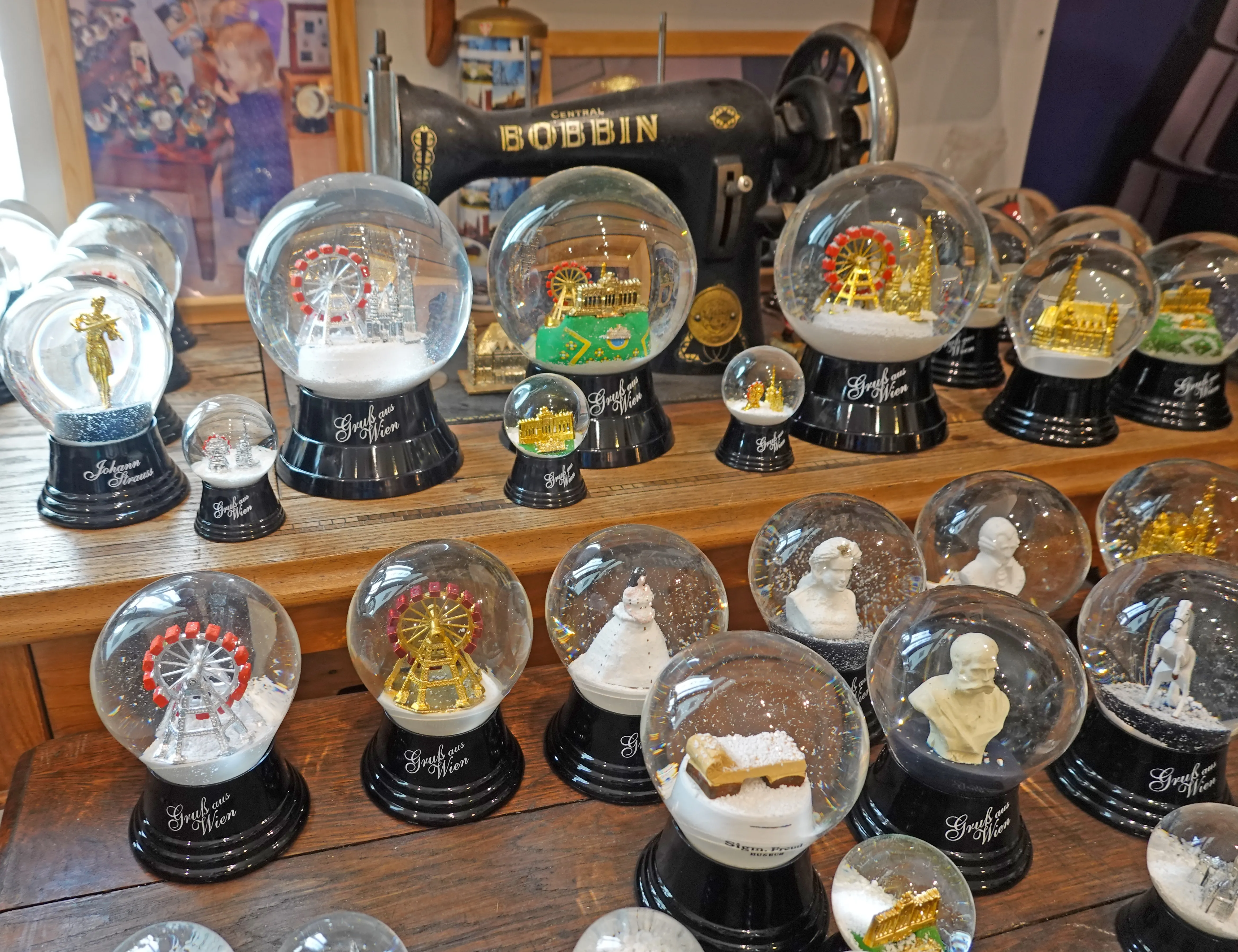 A collection of snow globes on display, featuring miniature scenes and figures, with a sewing machine behind them