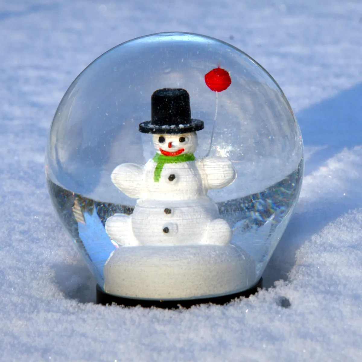 A snow globe with a snowman wearing a black hat and green scarf, holding a red balloon, placed in snow