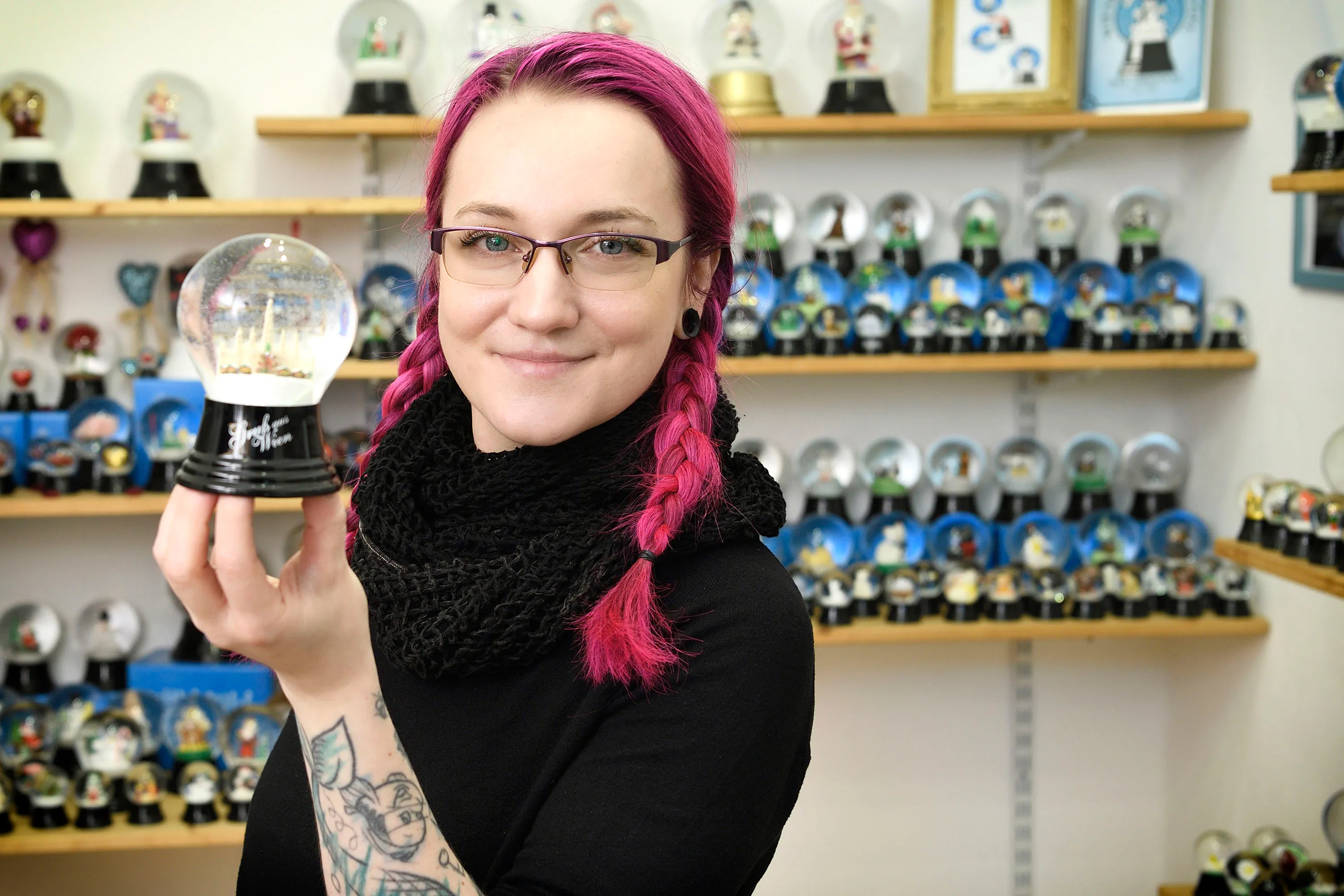 A woman with pink hair and glasses holds a snow globe, standing in front of shelves filled with snow globes.