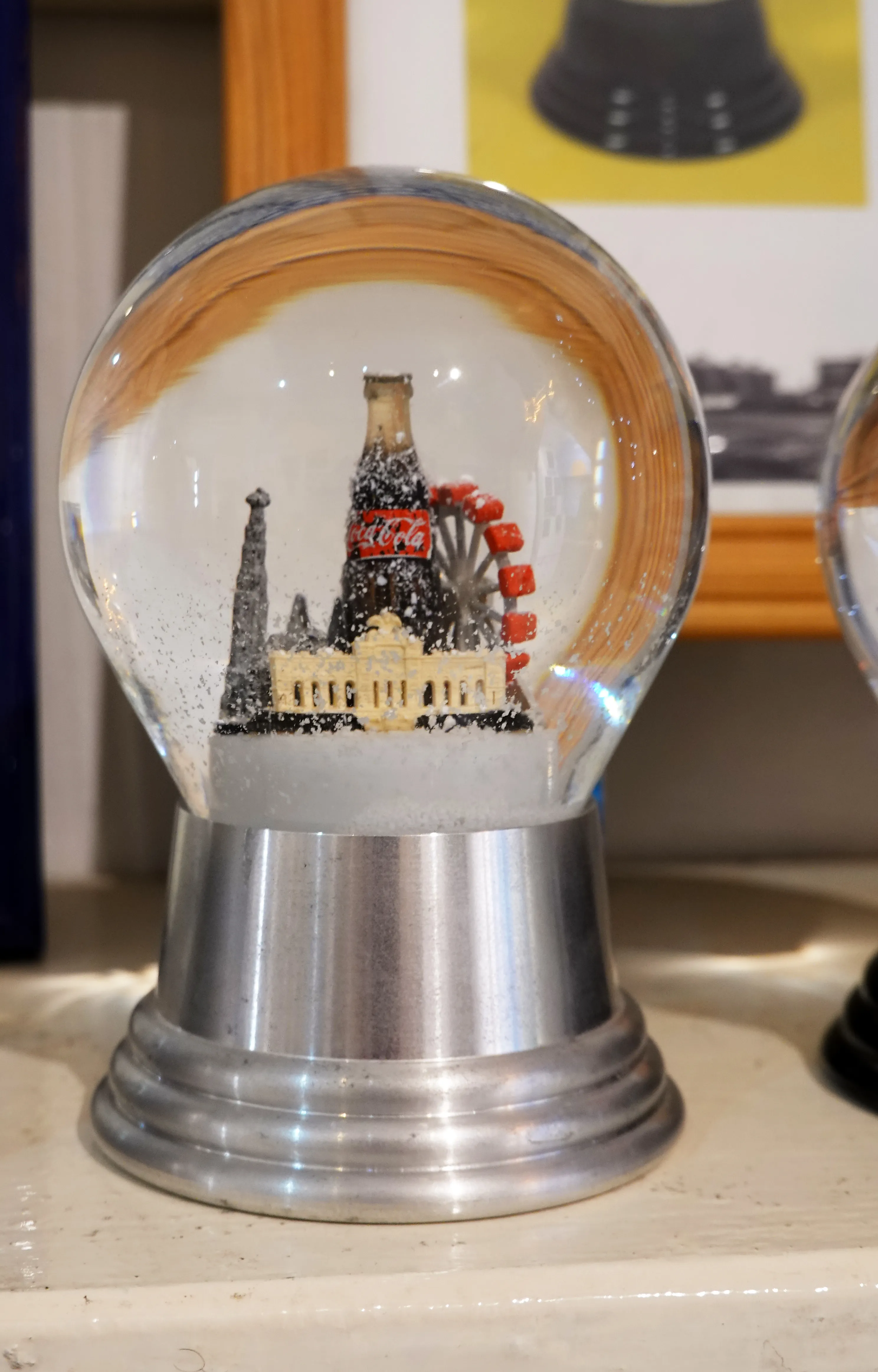 A snow globe with a miniature scene including a Coca-Cola bottle, a Ferris wheel, and buildings, displayed on a metallic base