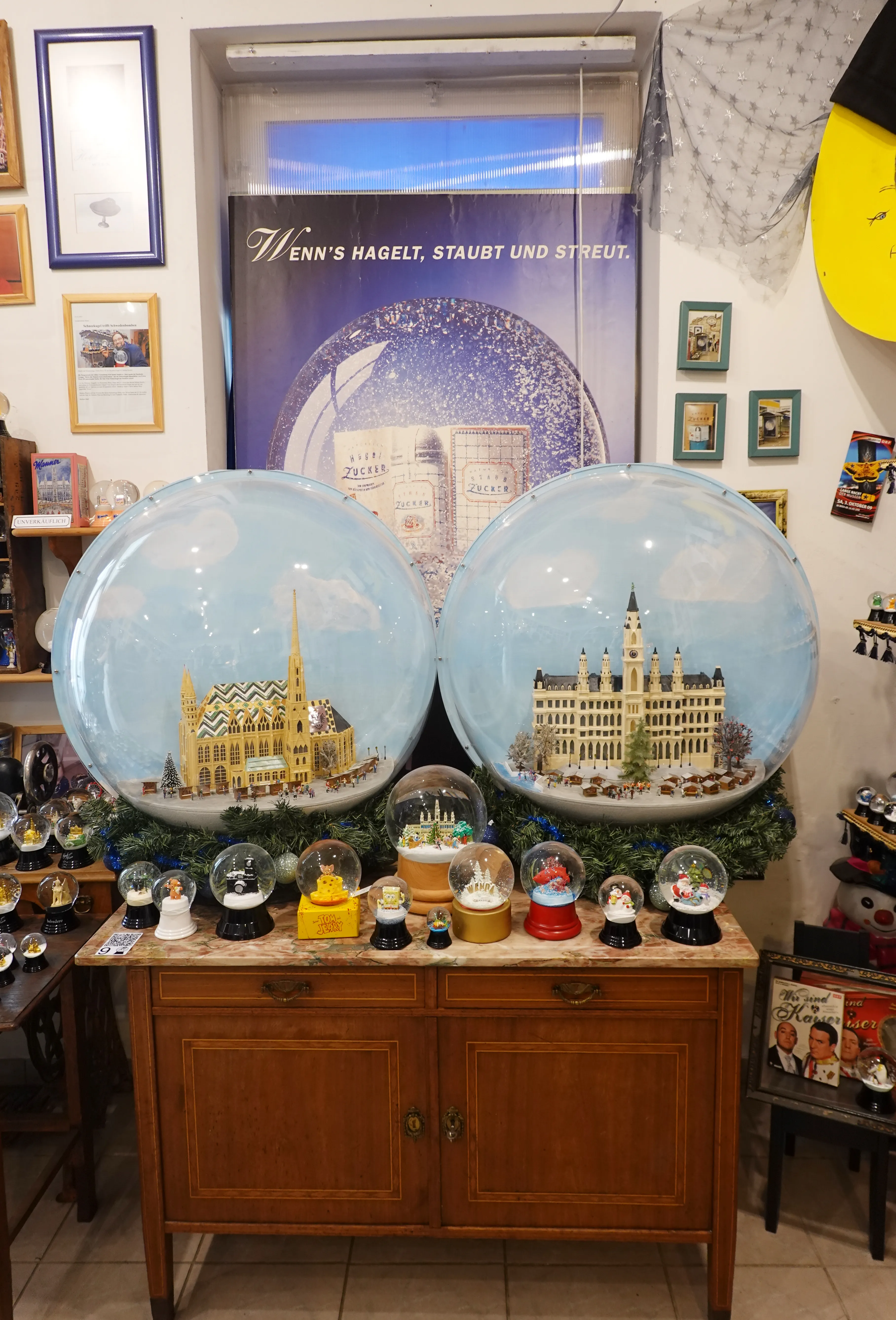 Dwie duże kule śnieżne z miniaturowymi budynkami w środku, ustawione na drewnianej szafce w muzealnej scenerii