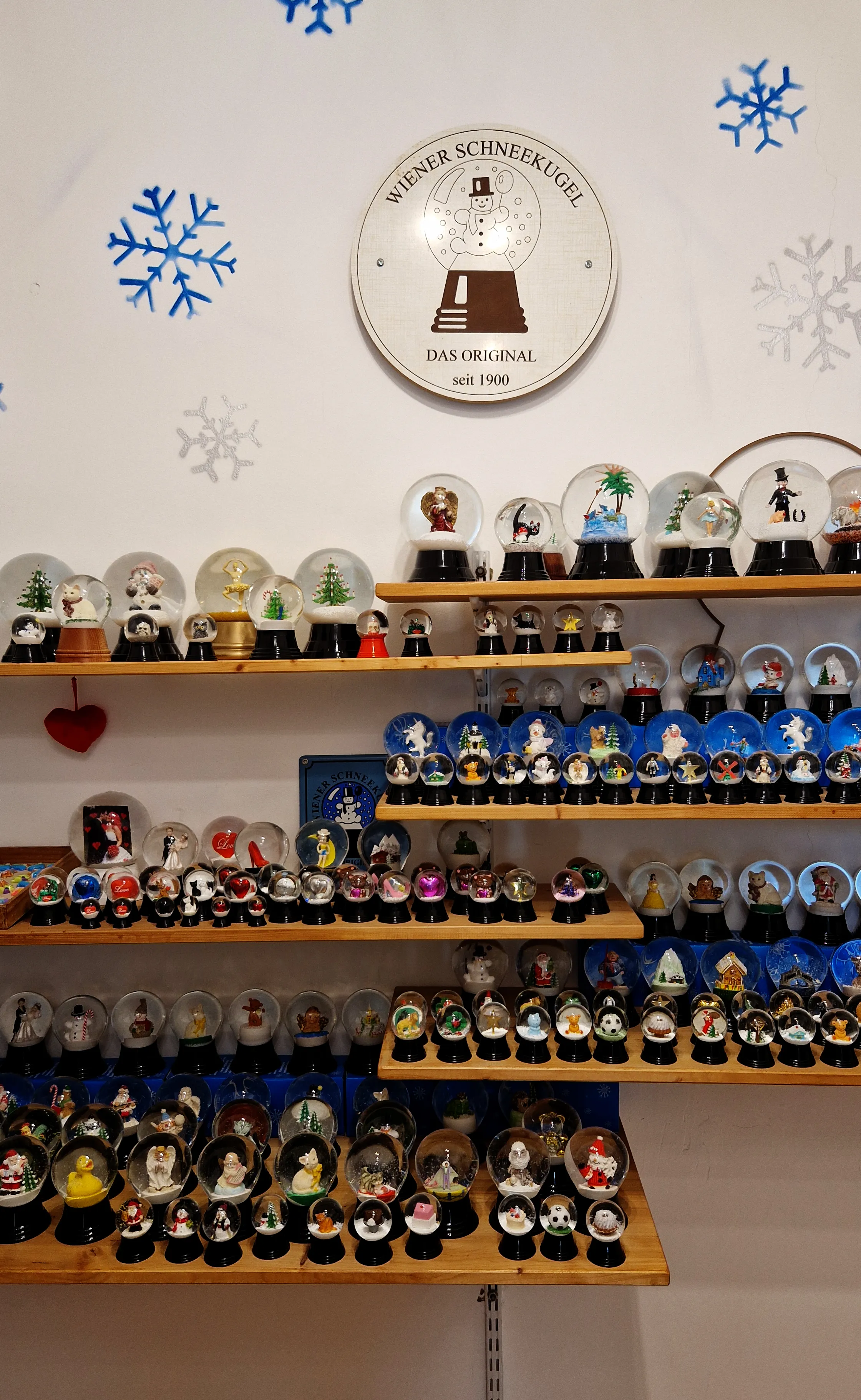 Shelves filled with numerous snow globes of various designs and sizes, with a circular sign on the wall above reading 'Wiener Schneekugel Das Original seit 1900' and blue snowflake decorations.