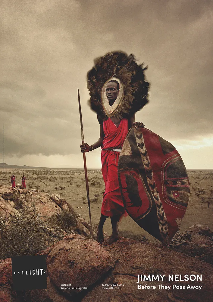 A Maasai warrior in red clothing and a feathered headdress holding a spear and shield, standing on rocks in a desert landscape under a cloudy sky.