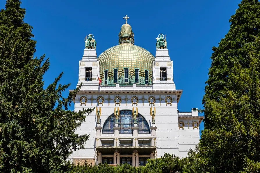 Kościół przy Steinhof ze złotą kopułą i fasadą z białego kamienia, w otoczeniu zielonych drzew pod błękitnym niebem