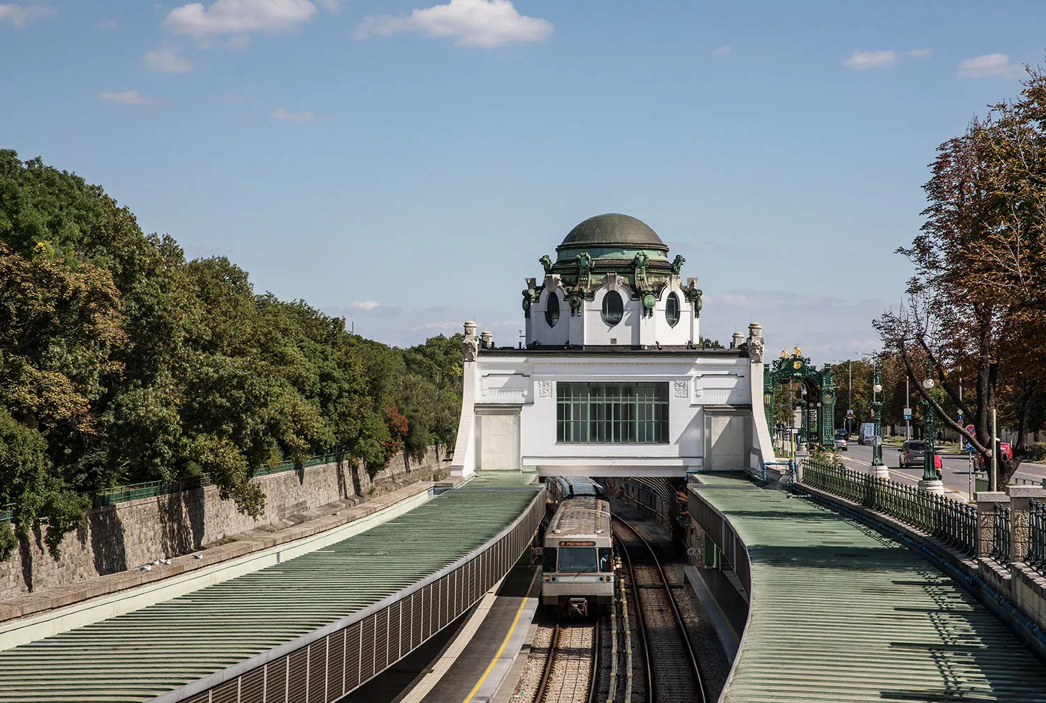 Een wit paviljoen met een groene koepel en sierlijke details staat boven een ingang van een treinstation, omringd door groene bomen en sporen onder een blauwe hemel.